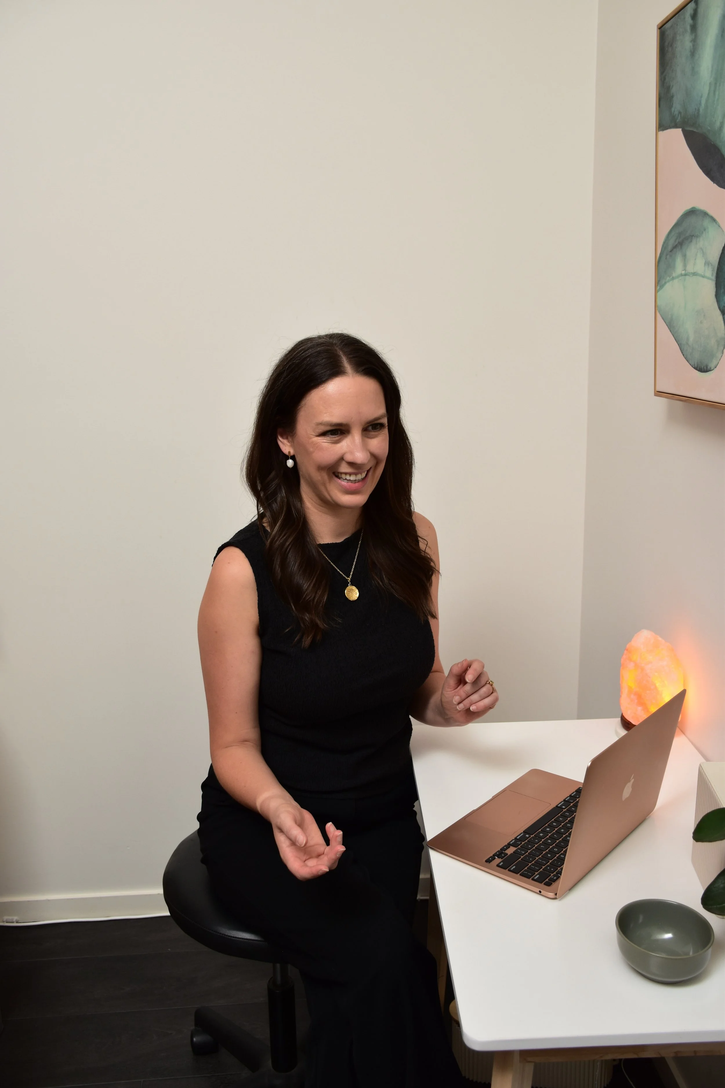 Alice Morgan, Acupuncture and Chinese Medicine practitioner, smiling during an online consultation at her desk with a rose gold laptop and salt lamp in a serene clinic setting