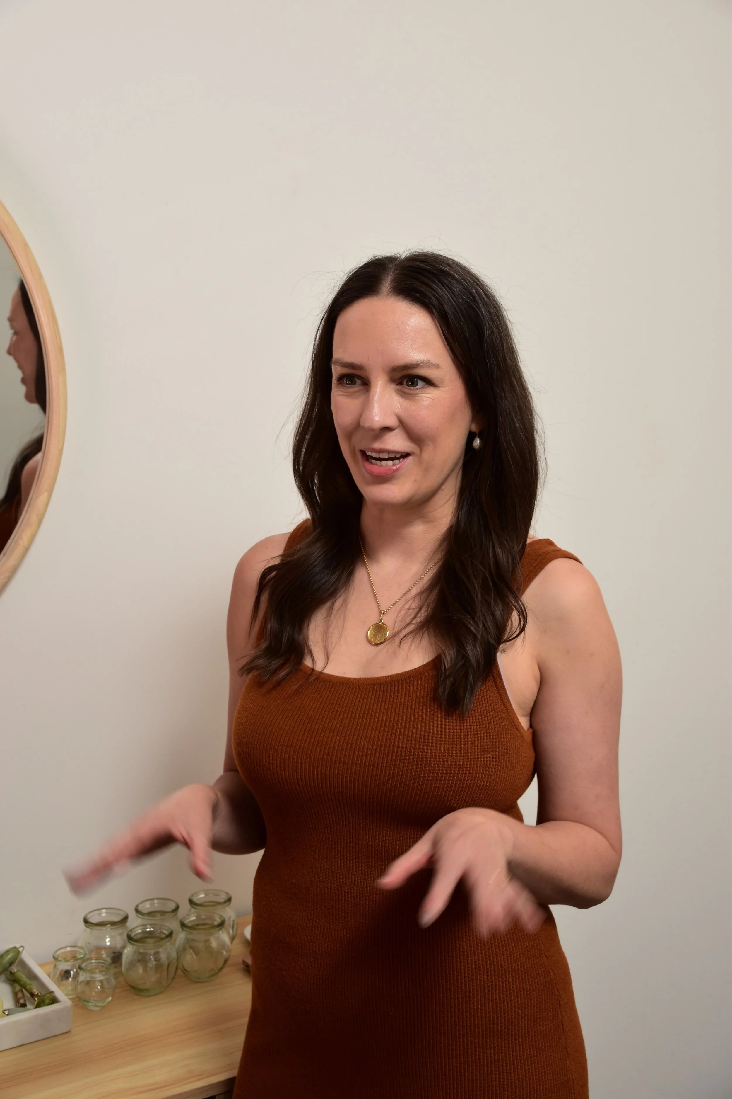 Portrait of Alice Morgan, a registered Acupuncture and Chinese Medicine practitioner, wearing a rust-coloured knit dress, gesturing warmly in a bright, modern clinic setting