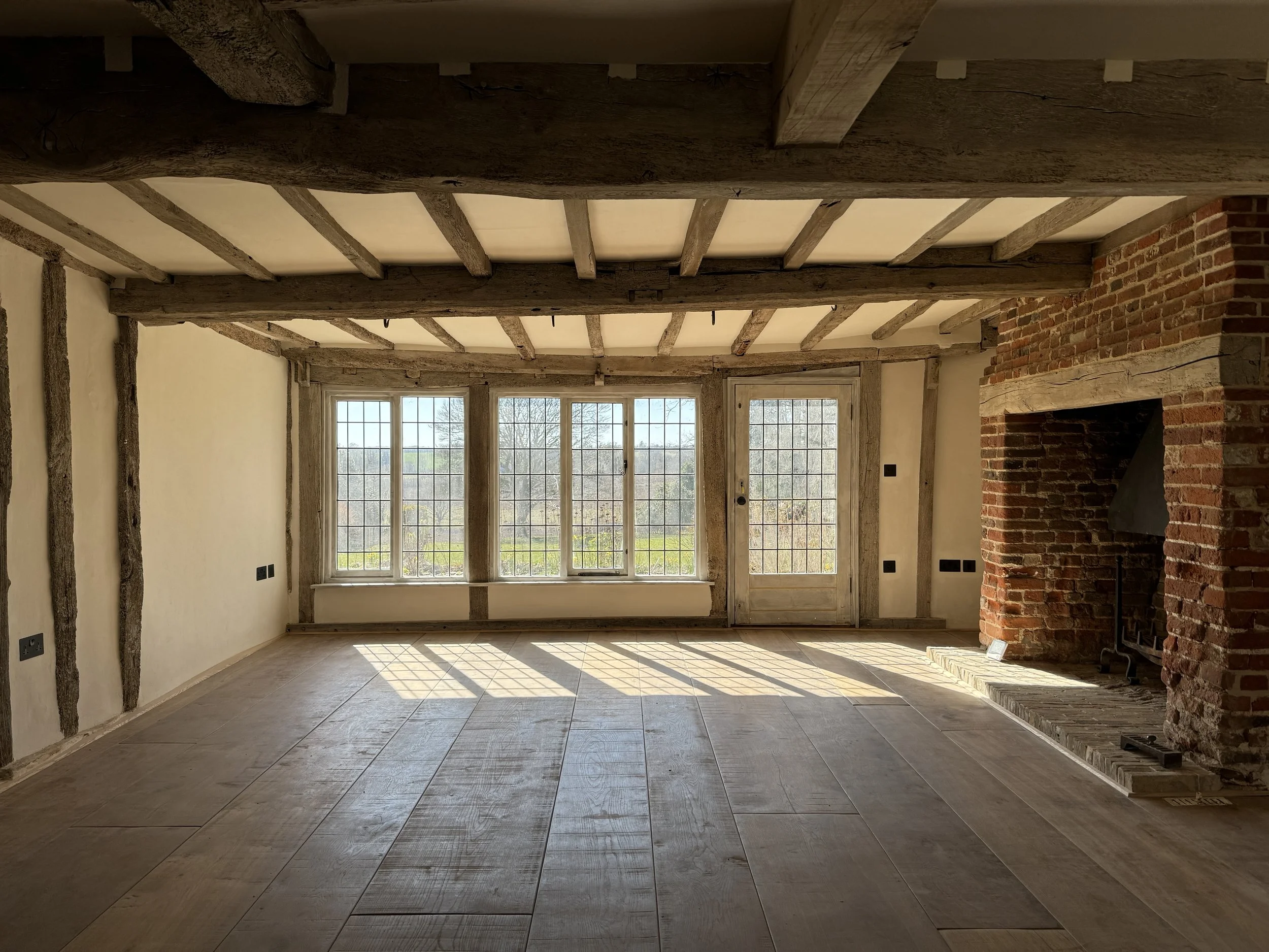 John Stebbing Architects Ltd — Empty room with large windows letting in natural light, exposed wooden beams on the ceiling, a brick fireplace on the right, and tiled flooring.