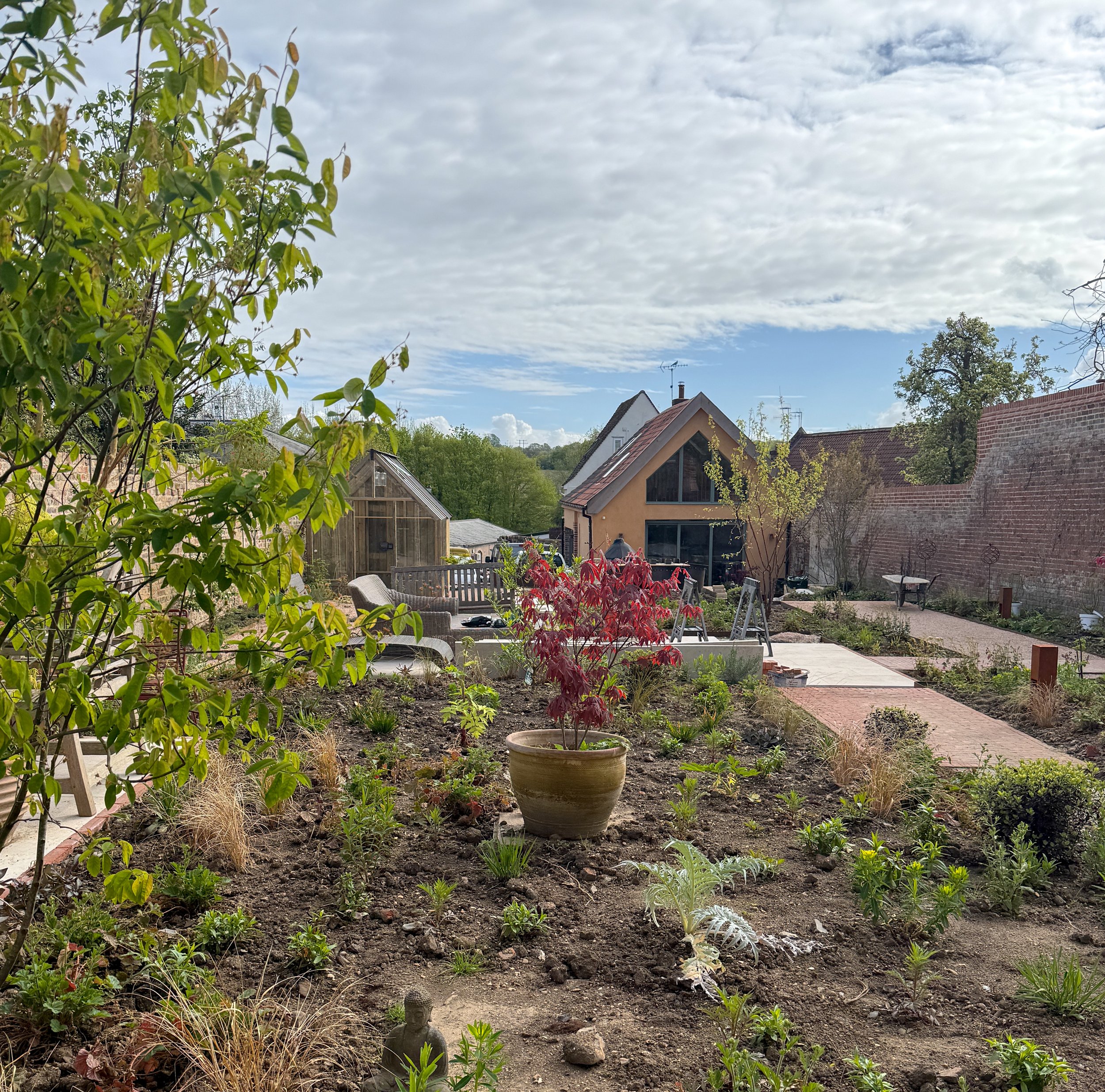 John Stebbing Architects Ltd — A backyard garden with various plants, a small Buddha statue, patio furniture, and houses with rooftops under a partly cloudy sky.
