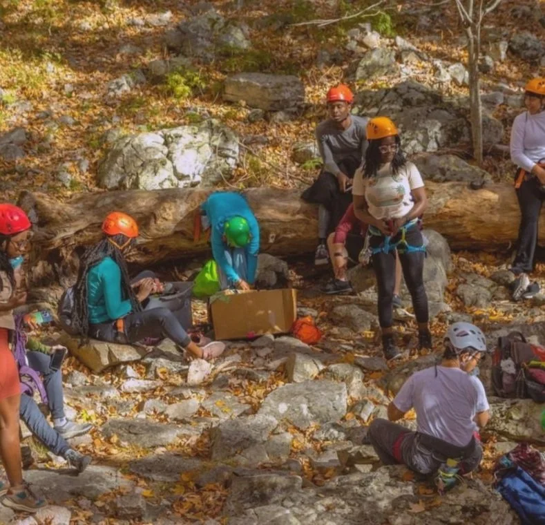 Group of people with helmets preparing for rock climbing or rappelling on a rocky, wooded outdoor area.