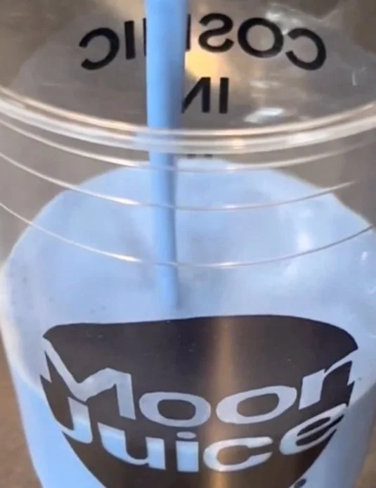 Close-up of a clear plastic cup with a straw, containing a blue-colored beverage, with the words 'Moon Juice' on the cup.
