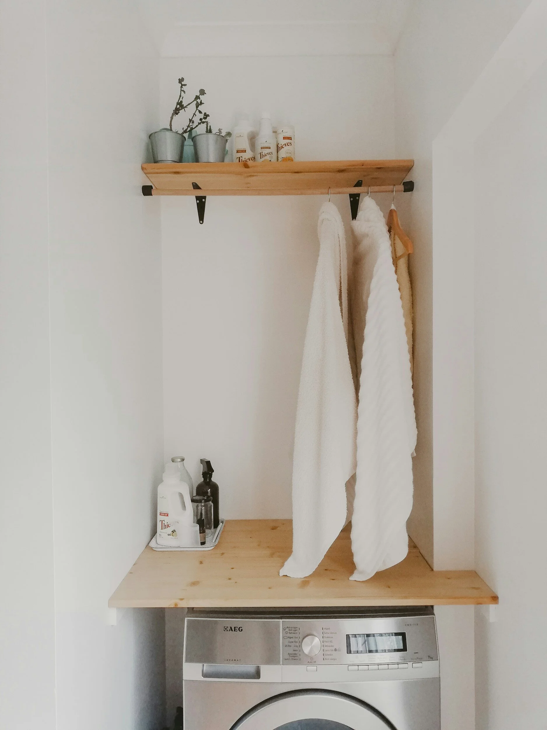 A laundry space with a wooden shelf and a hanging rod with two white towels and a wooden hanger. On top of the shelf, there are three small potted plants and bottles of Thieves cleaning products. Below, a wooden countertop is placed on top of a washing machine, which is partially visible.