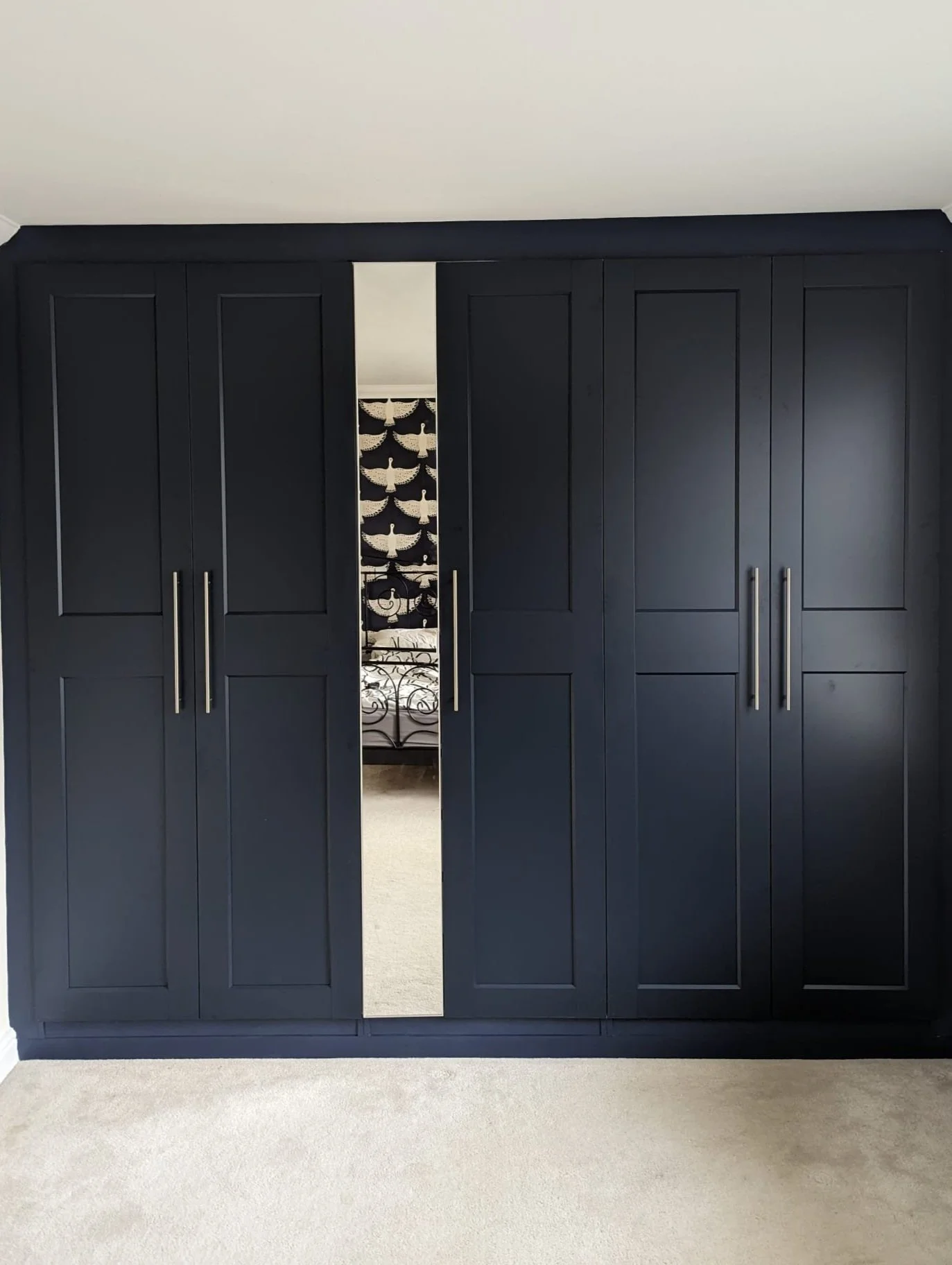 Black wardrobe with a mirror at the center reflecting a bedroom with a patterned wall and bed.