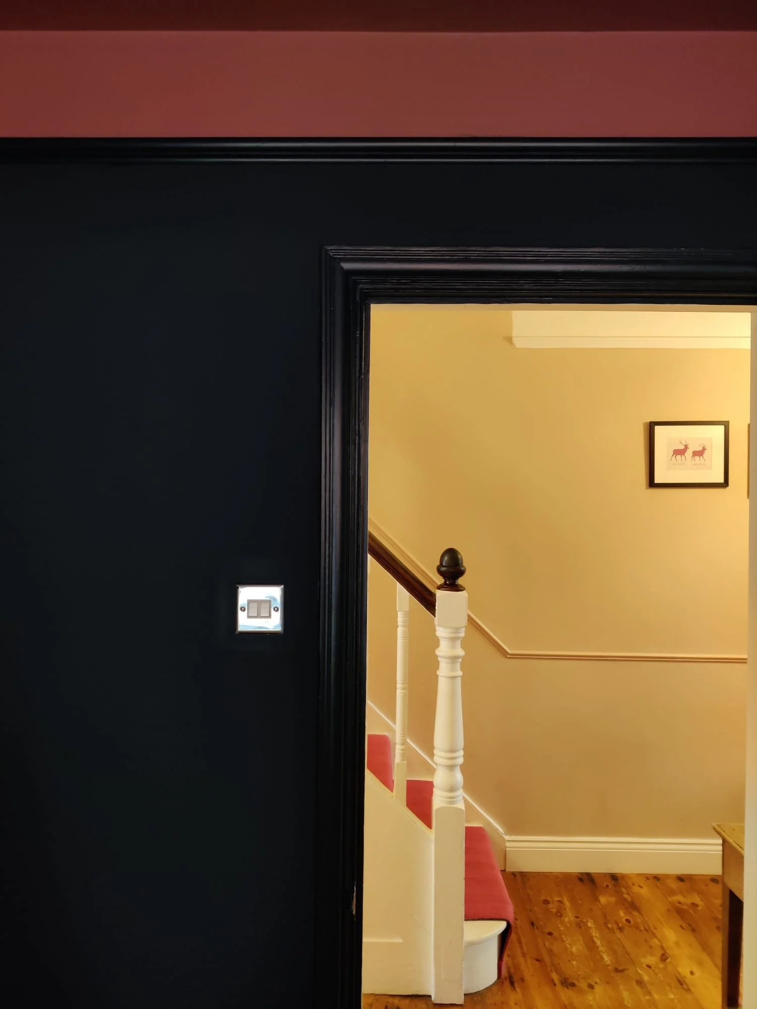 View of a hallway through a black door frame, showing a staircase with white balusters and a pink carpeted stair tread, cream-colored walls, wooden flooring, and a framed picture of two red animals on the wall.