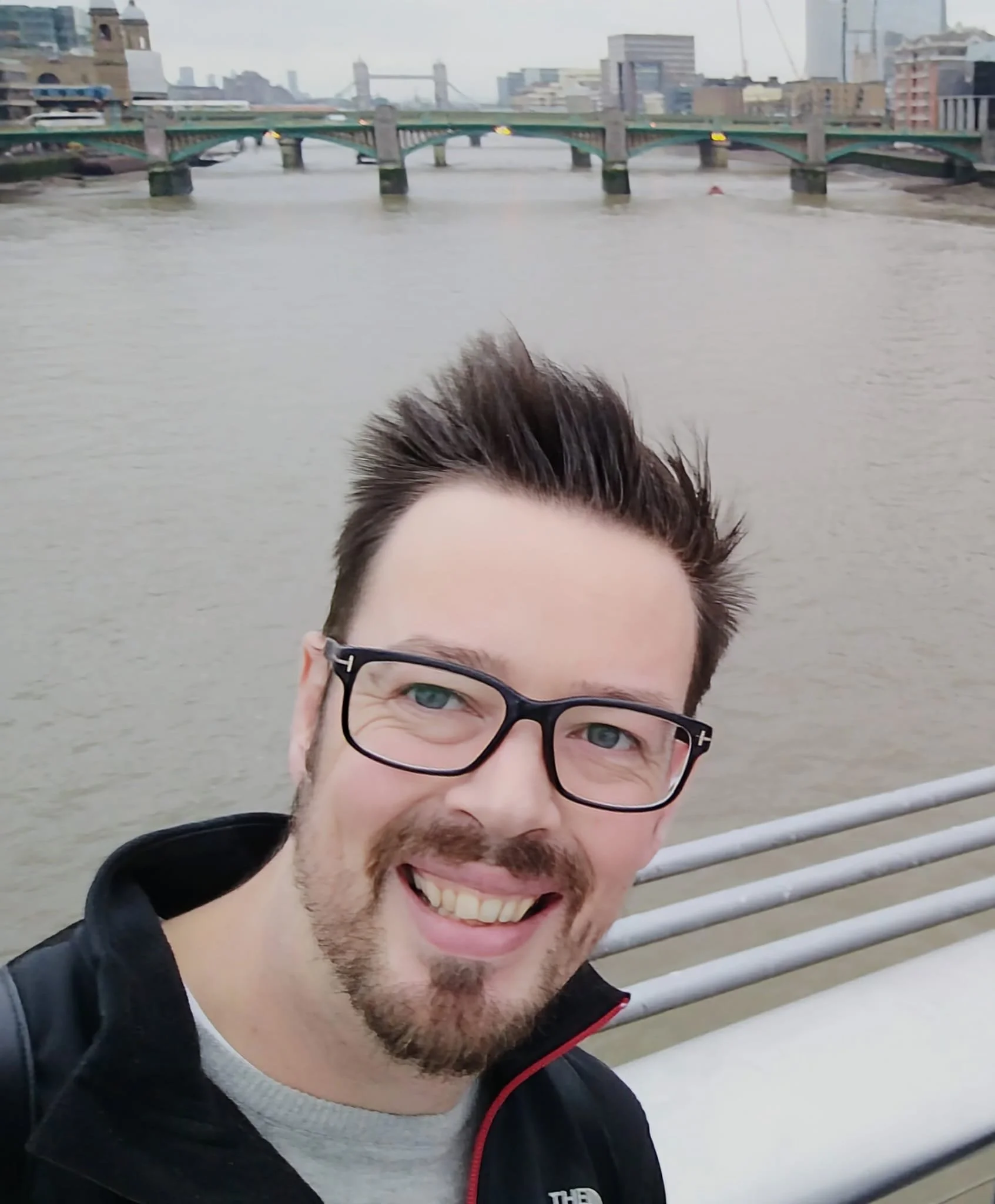 A smiling man with glasses and a beard taking a selfie on a bridge over the river with a city skyline in the background.