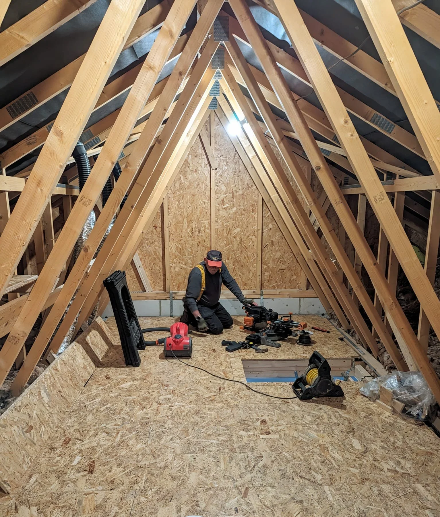 A person working inside an unfinished attic or loft space with exposed wooden beams, plywood flooring, and tools scattered around.