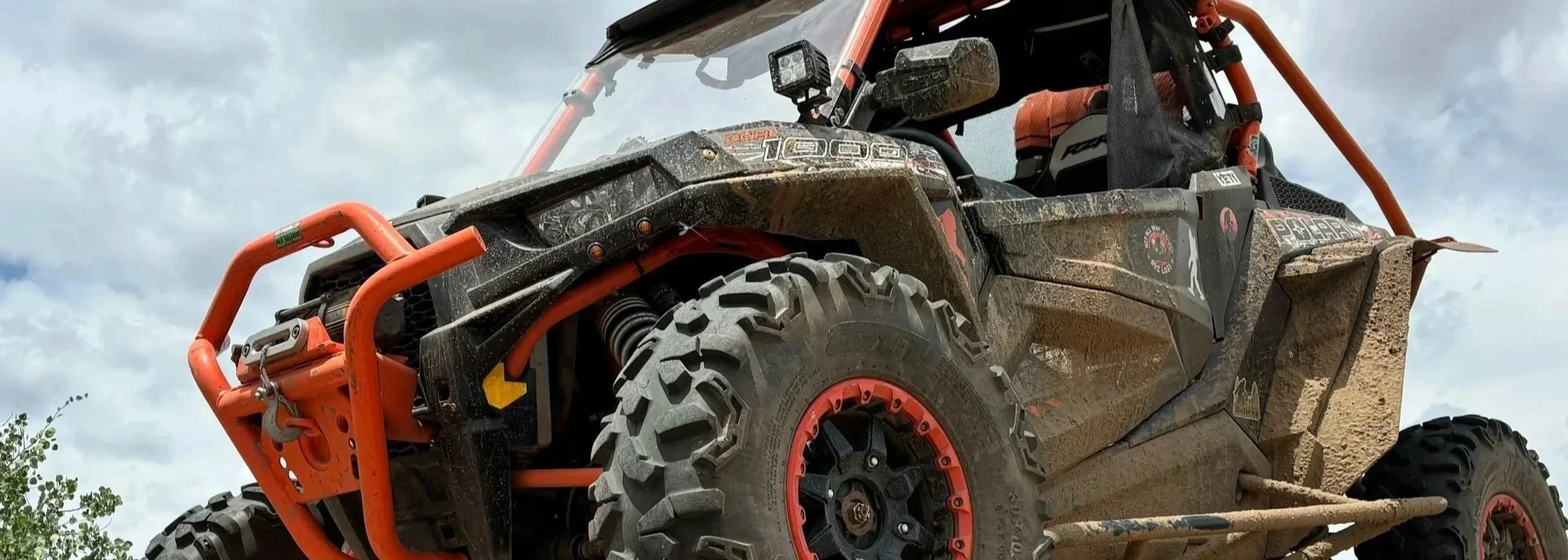 Off-road utility vehicle with large tires and orange roll cage, driving over dirt with mud splashes, under cloudy sky.