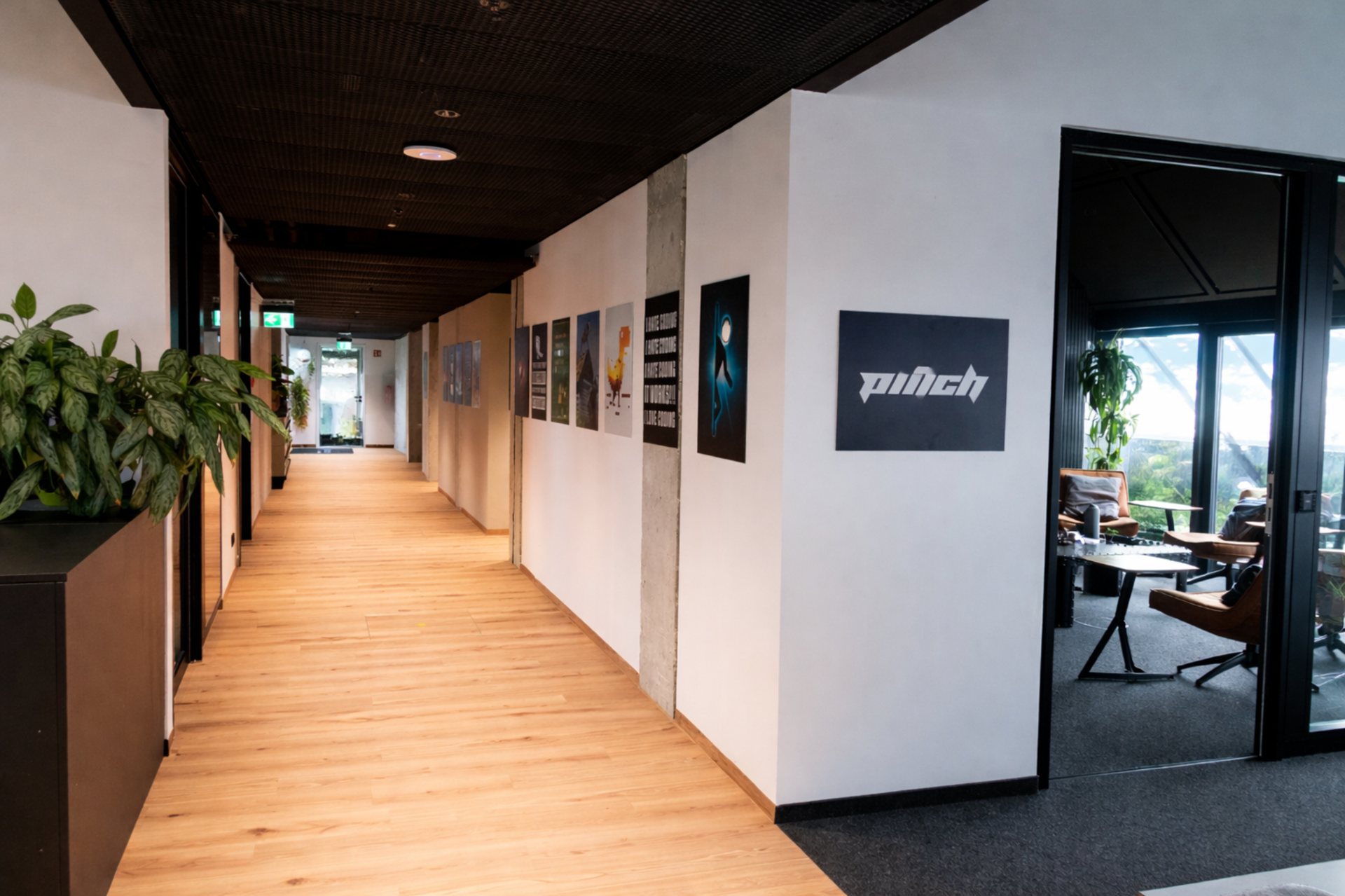 A hallway with wooden flooring, decorated with posters on the wall, leading to a door with windows. There is a large green plant on the left and a sitting area visible through a black-framed glass door on the right, with two people sitting and a view of an outdoor landscape.