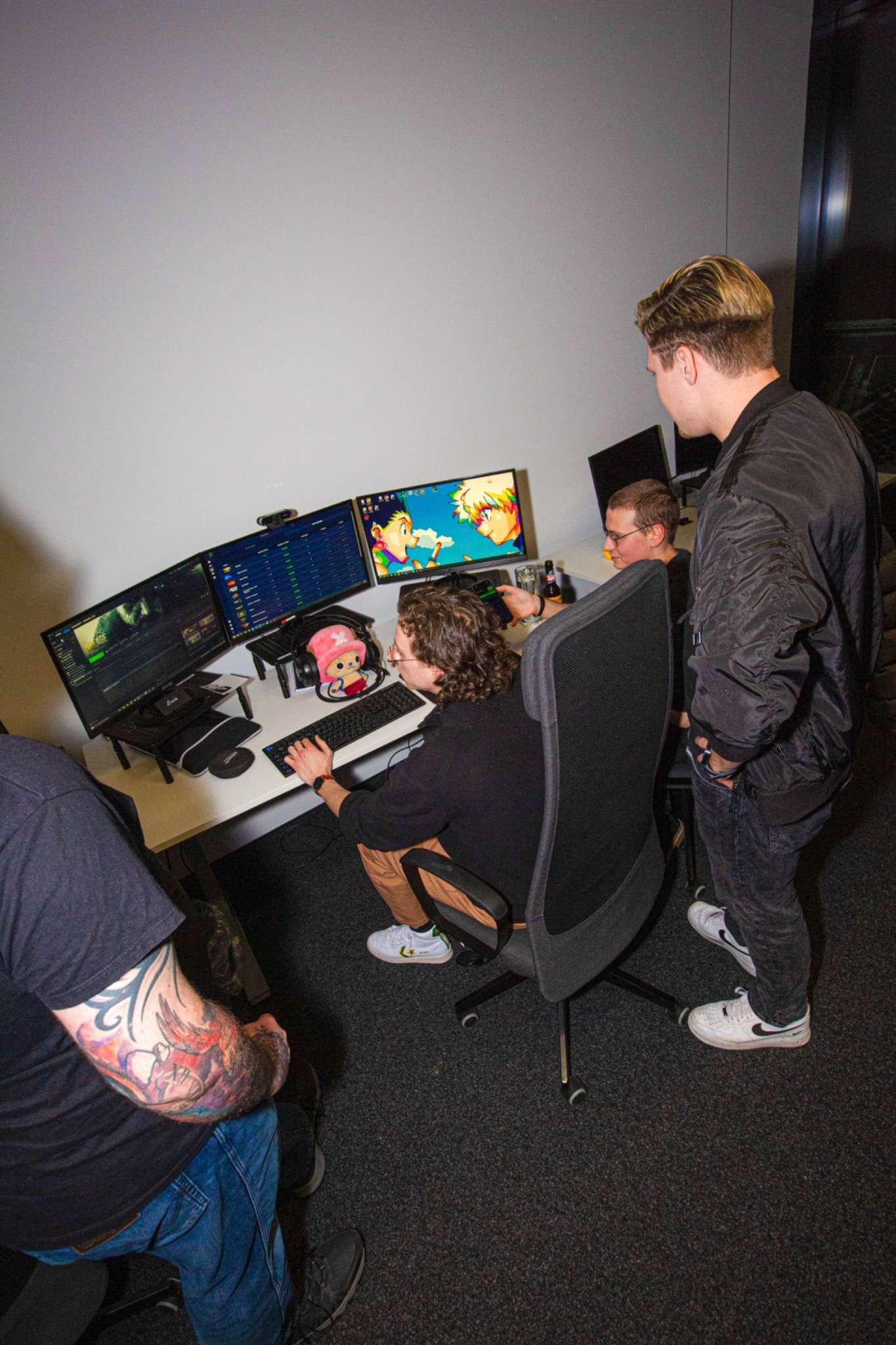 A group of people working at a desk with multiple computer monitors, two of which display anime-style characters. One person with glasses and curly hair is sitting and using a keyboard, while another person with tattoos on their arm stands nearby, observing. The desk has various items including a headset, a small figurine, and a drink. The room has a dark carpet and plain wall in the background.