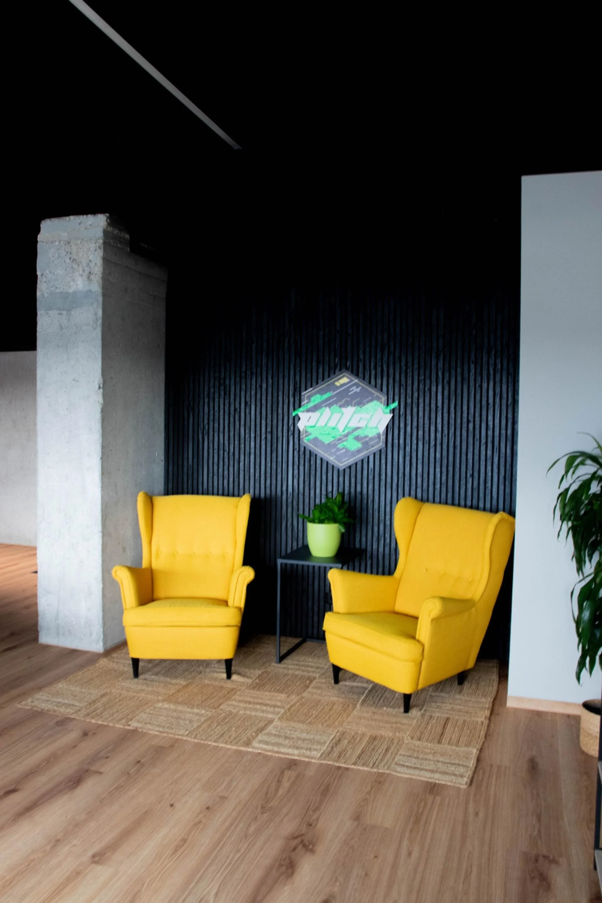 Two bright yellow armchairs sit side by side in front of a black panel wall, decorated with a small decorative table, a green potted plant, and a neon sign with Japanese characters. The room has wooden floors and a woven rug.