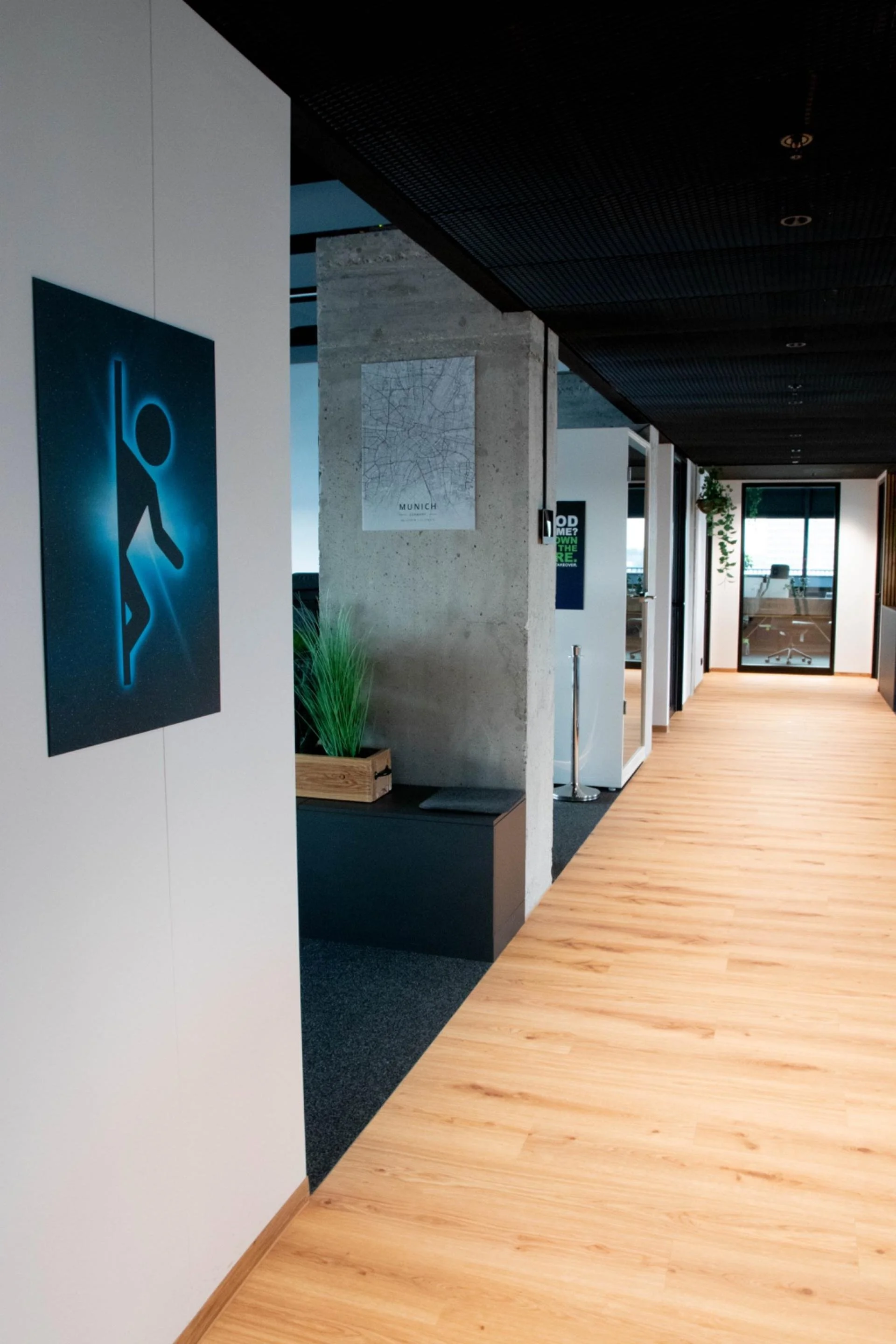 Modern office hallway with wooden flooring, white walls, and artwork, leading to a glass-walled office with a chair and desk in the background, illuminated by natural light from a large window.