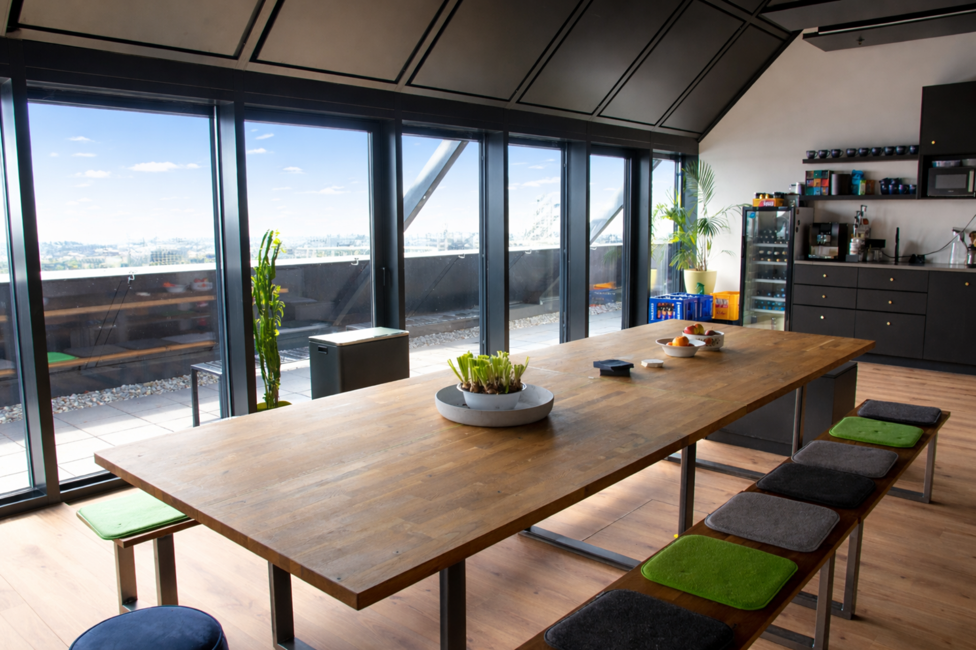 A modern kitchen with large floor-to-ceiling windows showing a cityscape view, a wooden dining table with bowls of fruit, potted plants, and a kitchen counter with appliances and shelves.