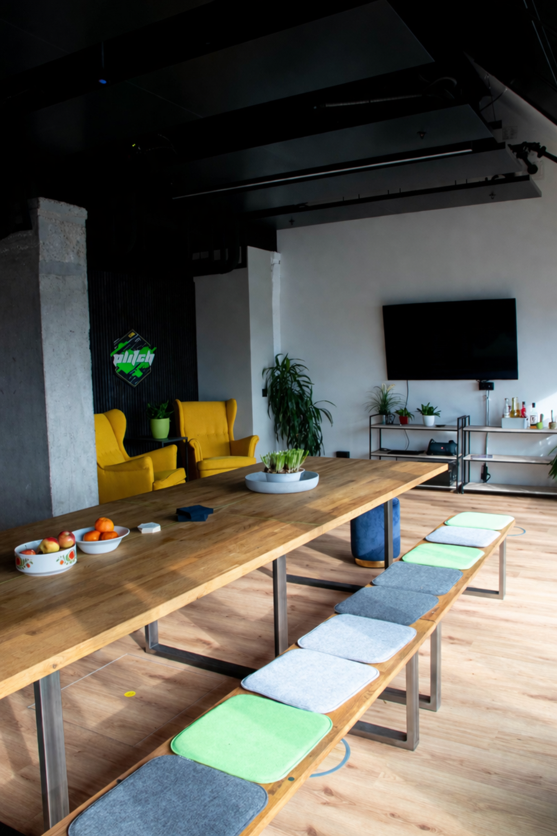 Modern office or conference room with a long wooden table, yellow armchair, and a flat-screen TV on the wall. Decor includes potted plants and a bowl of fruit on the table.