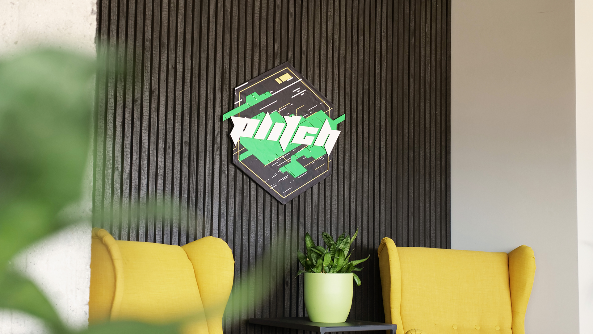 Interior lounge area with a black vertical slat wall, a decorative sign, yellow chairs, and a plant in a green pot.