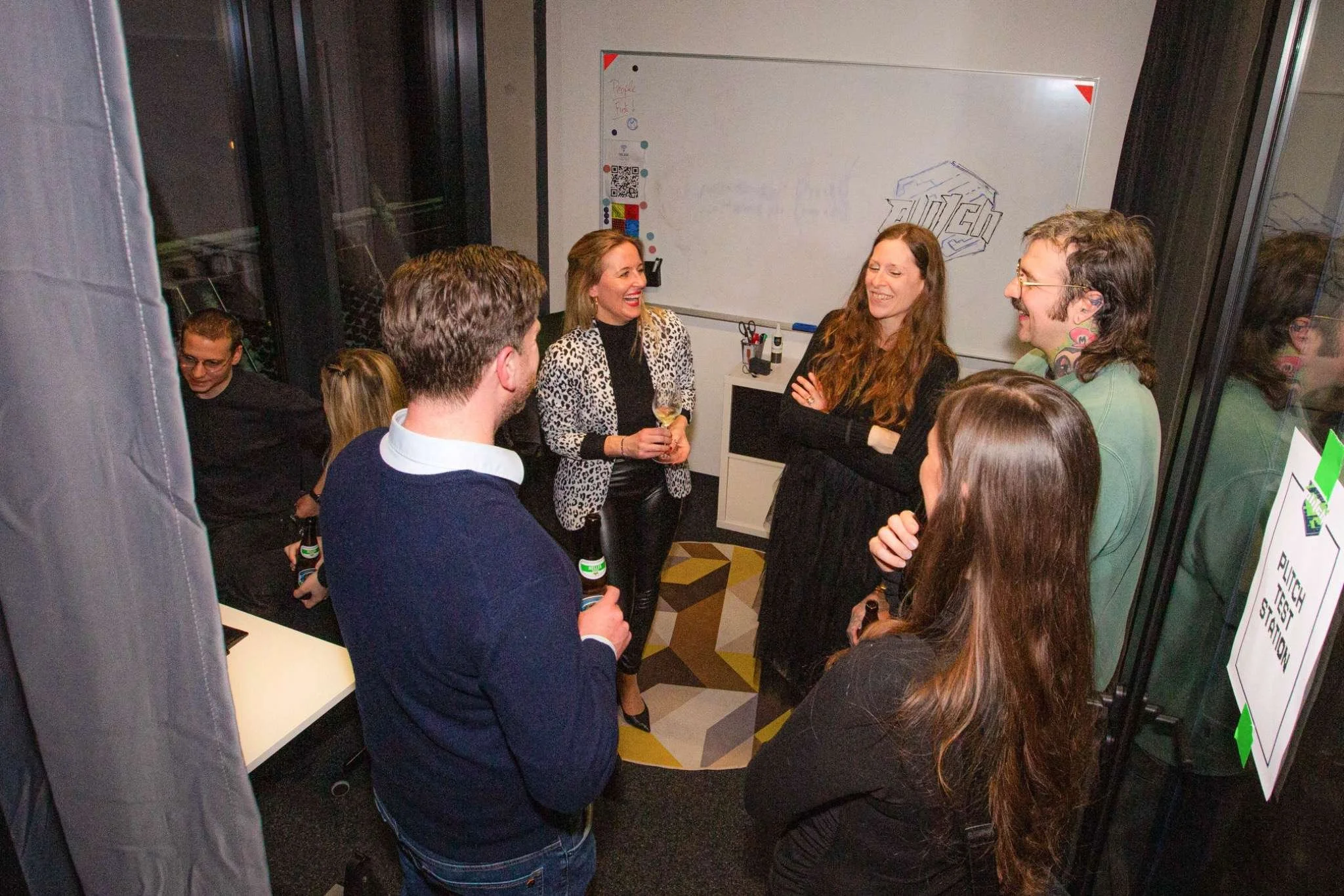 A group of seven adults gathered in an indoor space, engaging in conversation and smiling. Some are holding bottles, and a woman with a leopard print blazer is holding a glass of wine. There is a whiteboard in the background with drawings and text, a