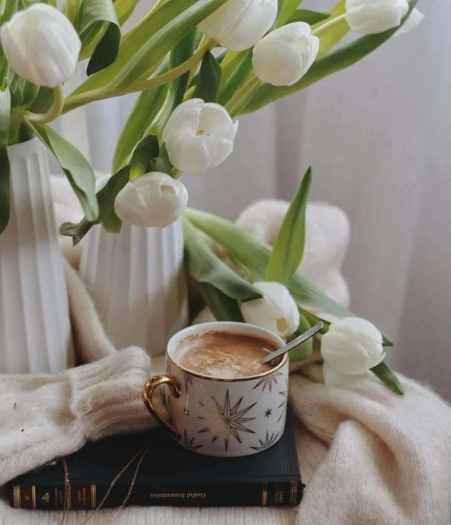 A table with a cup of hot chocolate and a vase with white tulips.