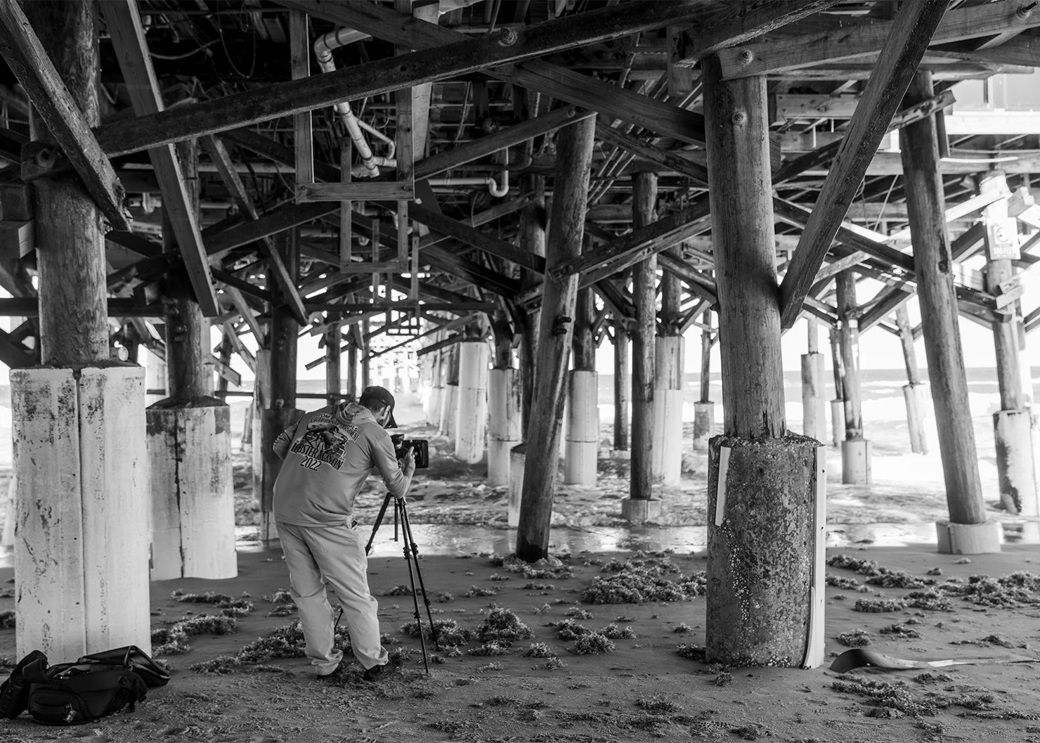 Fine Art photographer Bradley alan, under a pier, taking a long exposure photo of the oceans waves slamming into the pier beams and the sand.