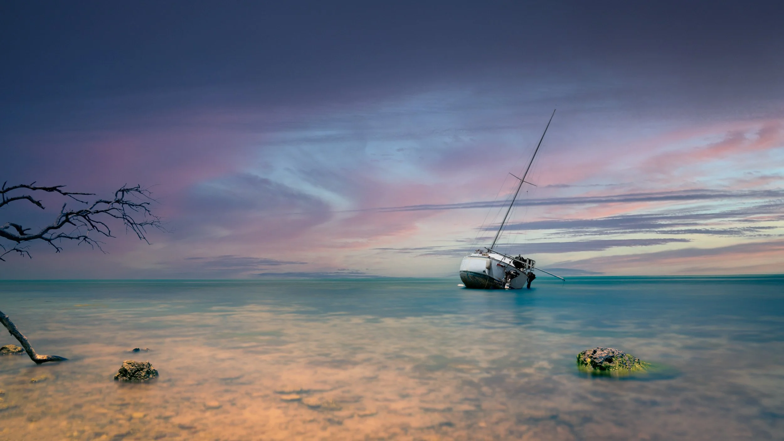 A boat partially submerged in calm water near rocks and a leafless tree branch, with a colorful sunset sky in the background. Minimalist long exposure fine art - Bradley Alan - Florida Keys - limited edition print