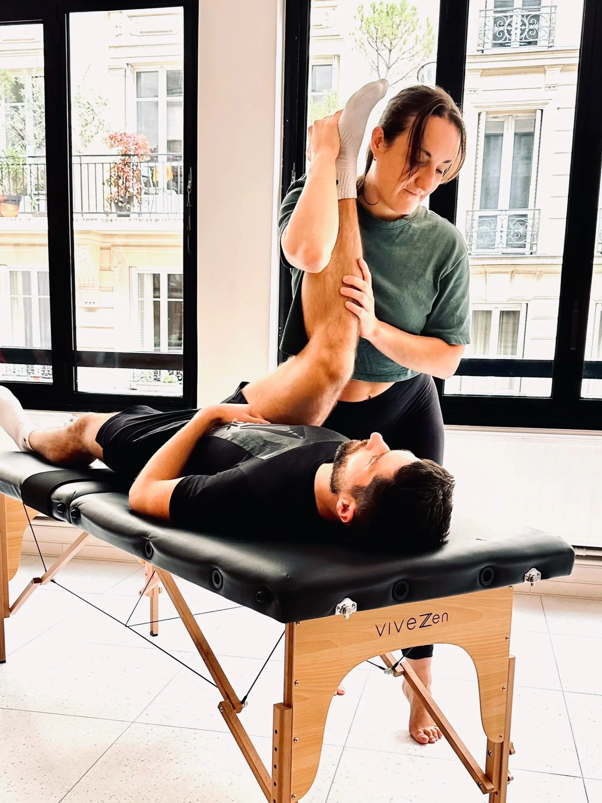 Une femme effectue une thérapie de rééducation avec un homme allongé sur une table de physiothérapie, dans une pièce lumineuse avec de grandes fenêtres.