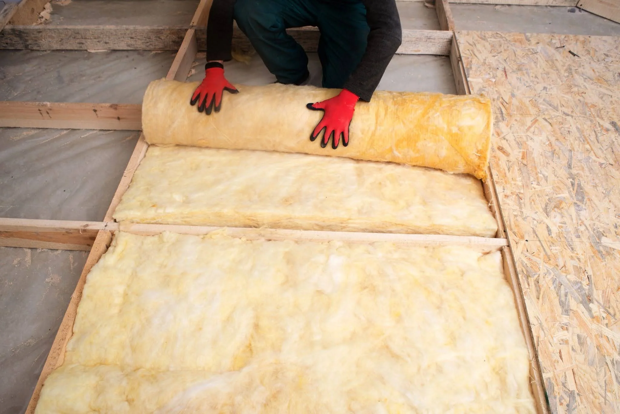 Overhead view of a worker wearing red gloves unrolling a long roll of yellow unfaced fiberglass insulation between wooden floor joists.