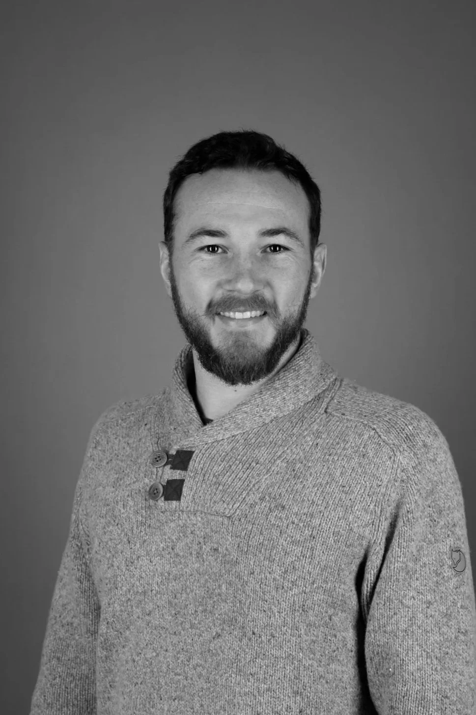 Black and white photo of a young man with curly hair, smiling, wearing a white button-up shirt with chest pockets, against a plain background.