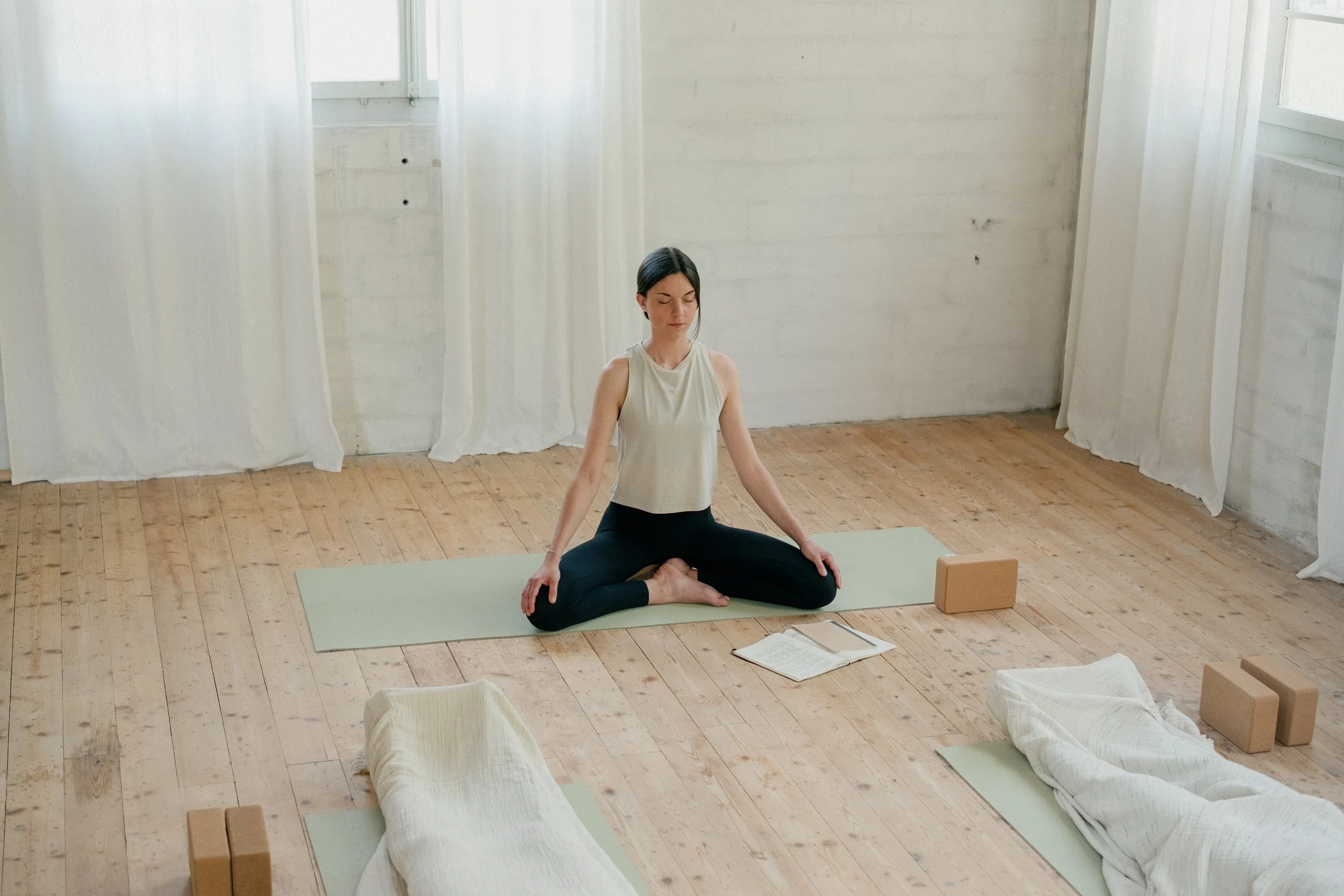 Eine Frau sitzt in einer meditativen Haltung auf einer Yogamatte in einem lichtdurchfluteten Yogastudio in Konstanz. Ein achtsamer Moment der Entspannung