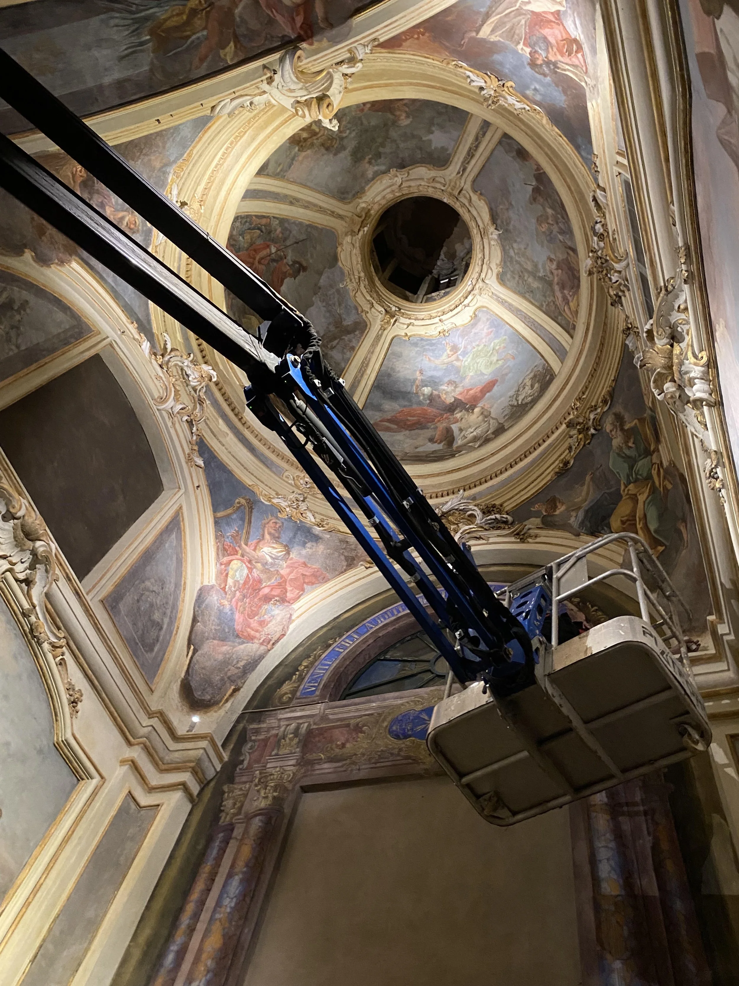 Un lift sta portando un operatore verso il soffitto di una chiesa o un edificio storico che ha affreschi e decorazioni barocche.