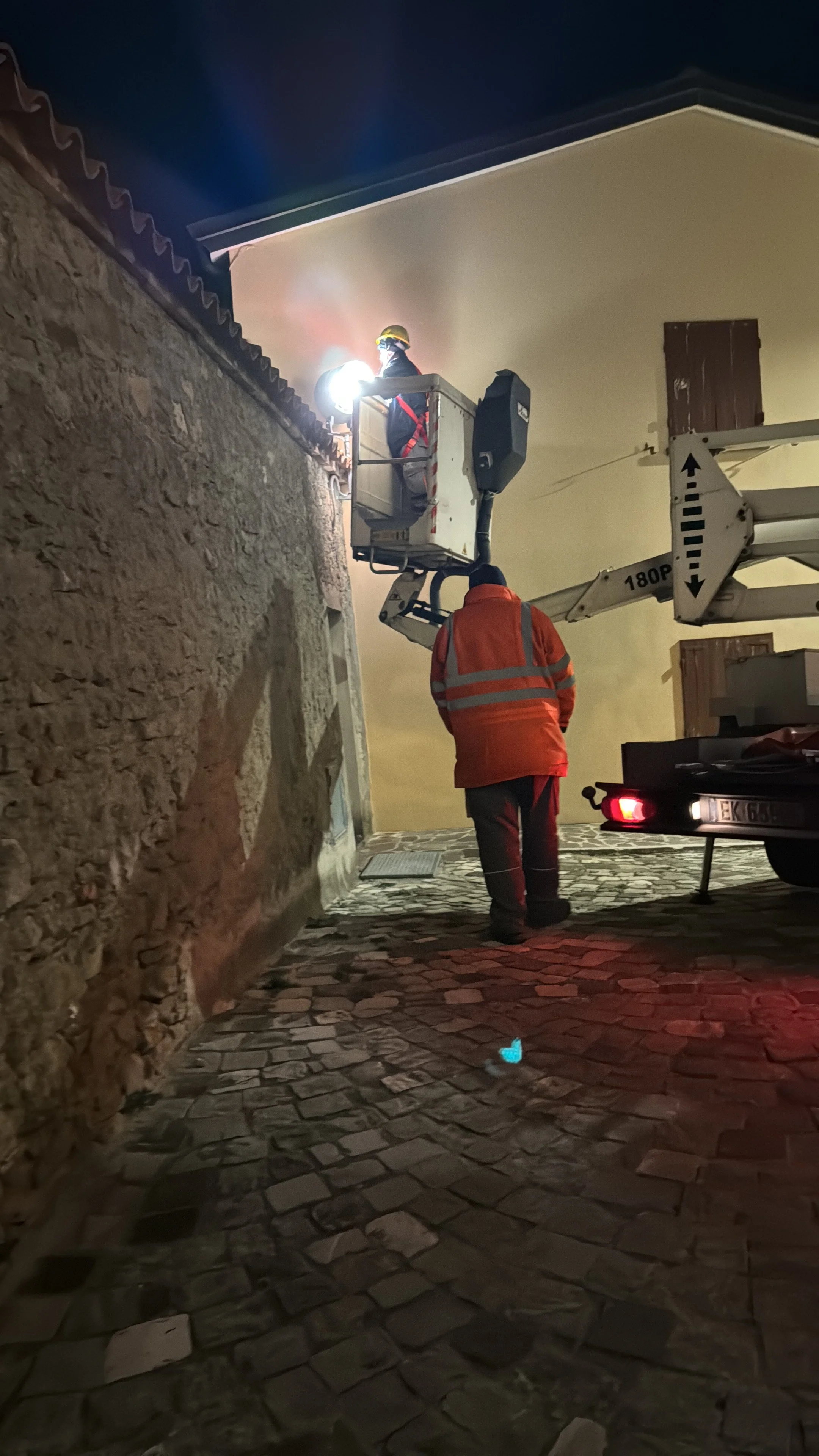 L'immagine mostra un intervento di un tecnico di emergenza antincendio che utilizza una piattaforma elevatrice per spegnere un incendio sul lato di una casa in una strada in pietra. È notte e ci sono due persone visibili, entrambe con indumenti ad alta visibilità e caschi.
