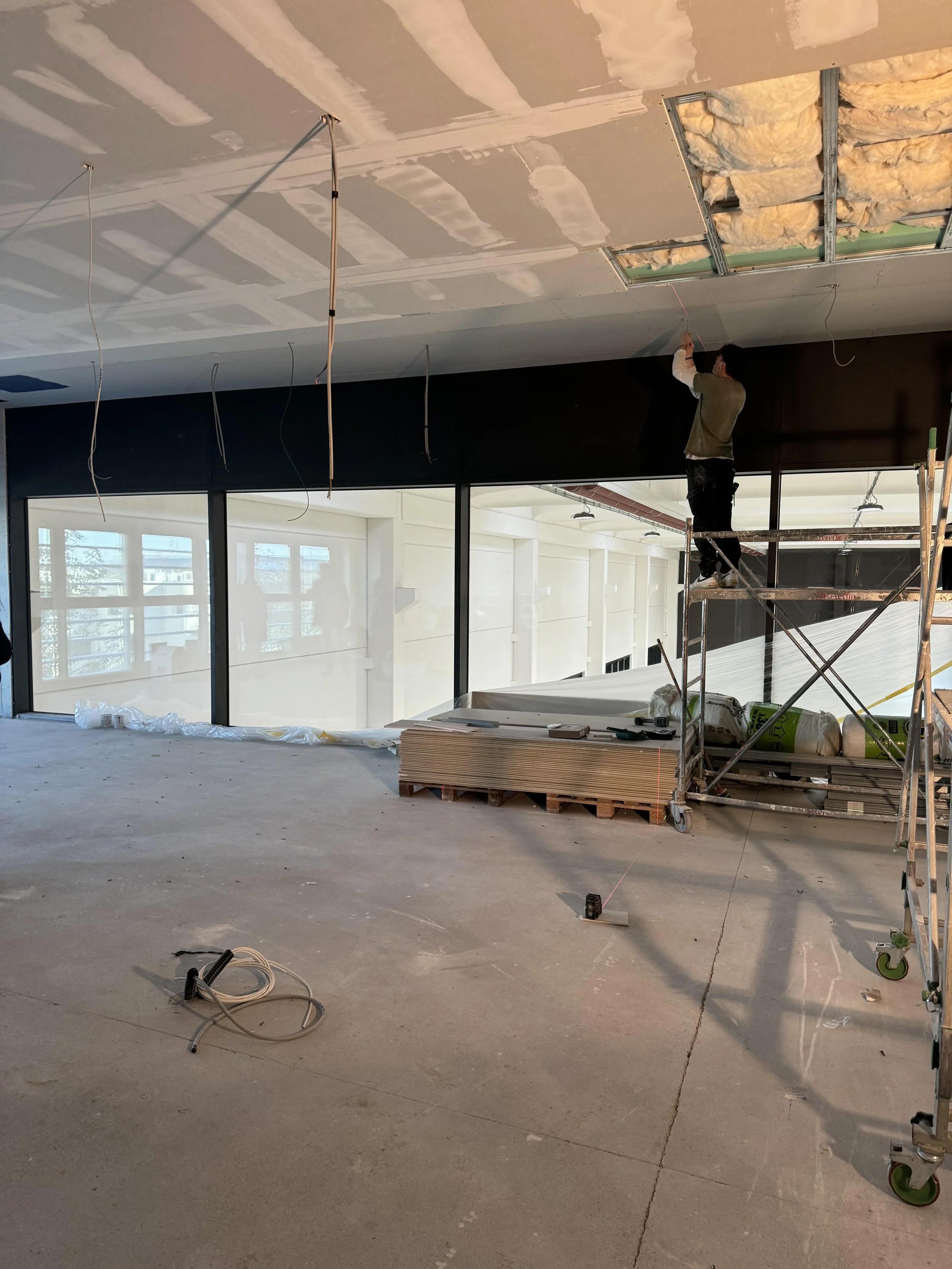 Lavoratore su un'impalcatura che installa o sistema controsoffitto in un ambiente interno di ufficio o commerciale, con finestre e luci a soffitto.