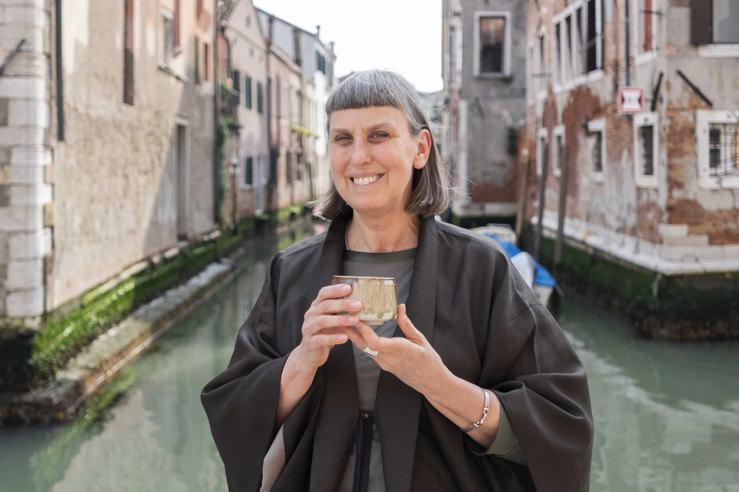 Donna con capelli corti e grigi sorridente che tiene una tazza mentre si trova in un'area urbana vicino a un canale o un fiume con edifici storici sullo sfondo.