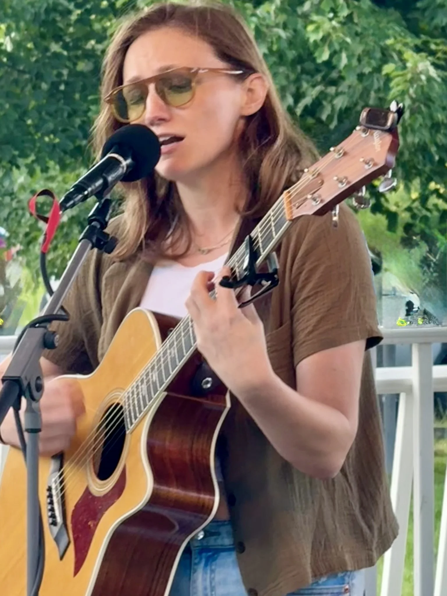 A woman wearing sunglasses playing an acoustic guitar and singing into a microphone outdoors with green trees in the background.