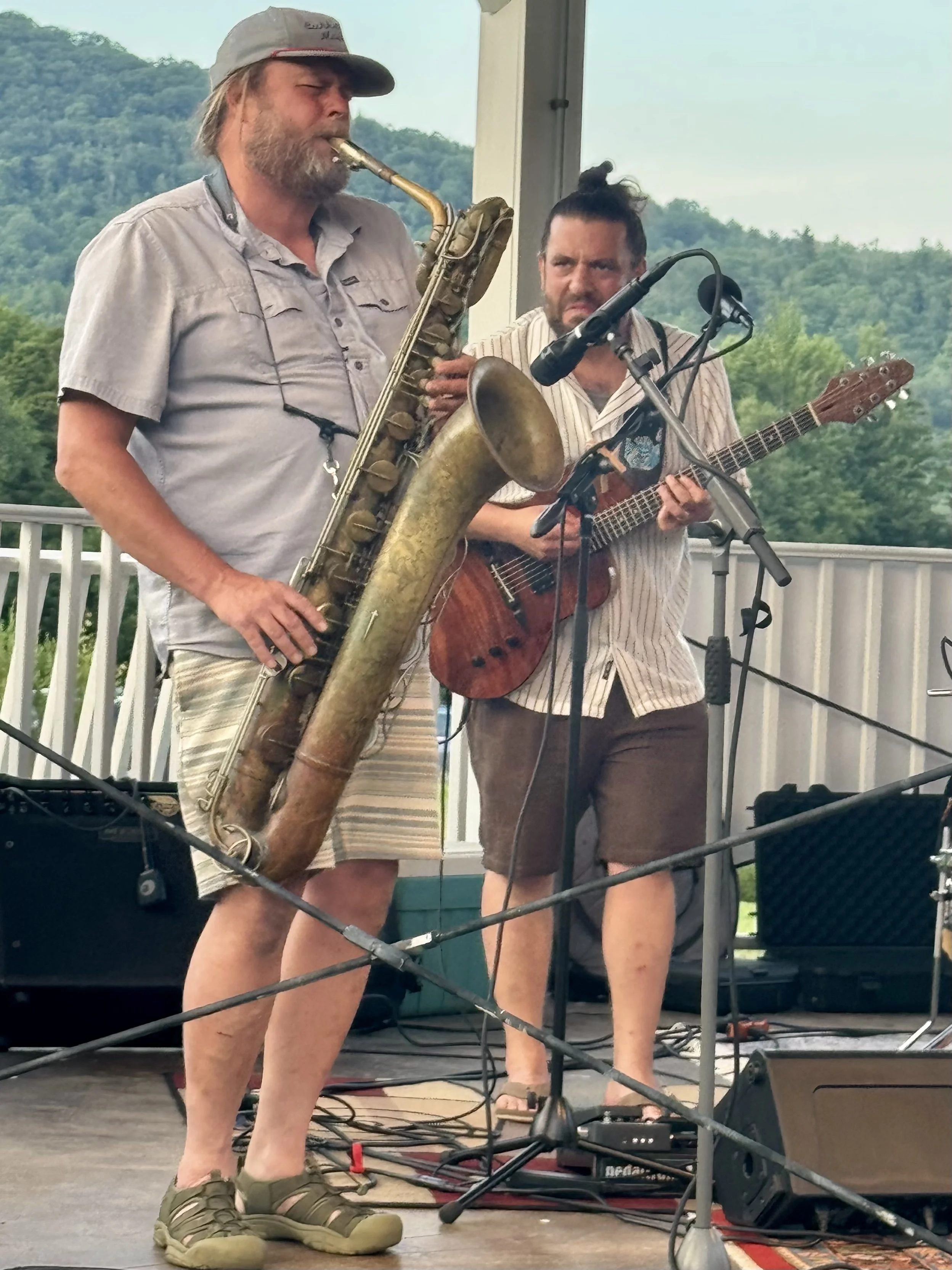 Two men performing on a stage with musical instruments; one playing a baritone saxophone, the other playing an electric guitar.