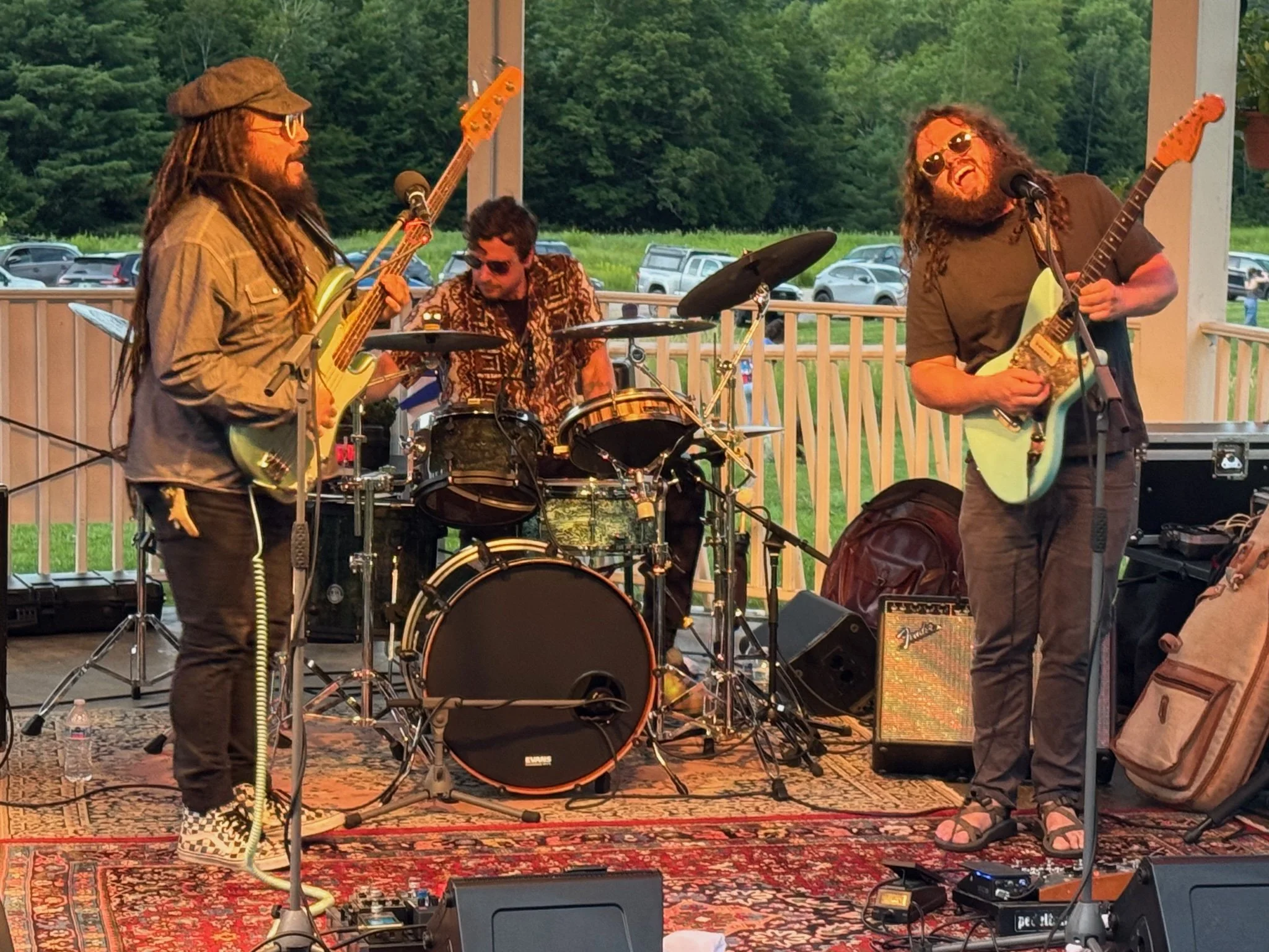 A band performing on an outdoor stage with three members playing guitars and drums, with a background of green trees and parked cars.