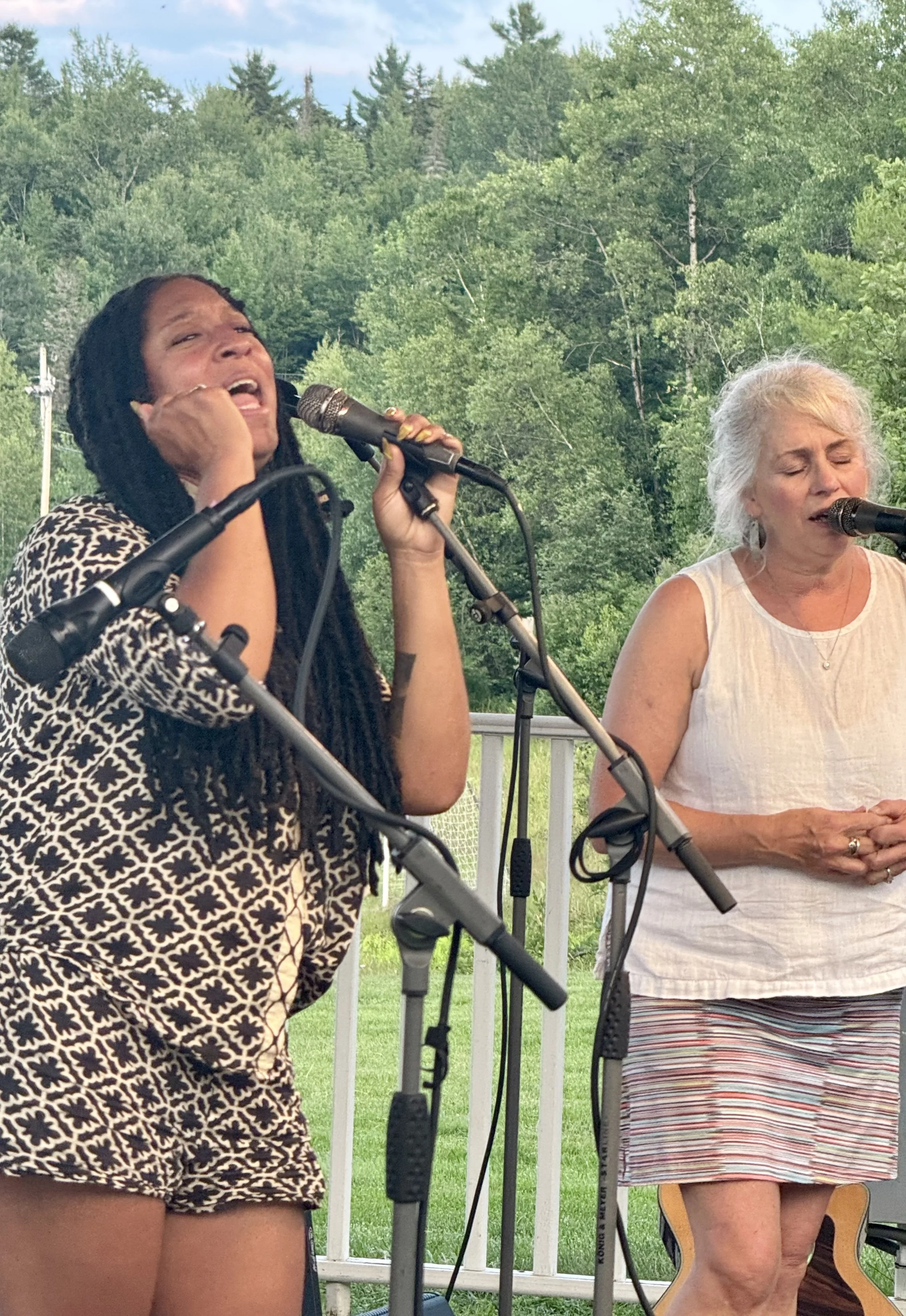 Two women singing in an outdoor setting with a green forest background, microphones, and musical equipment.