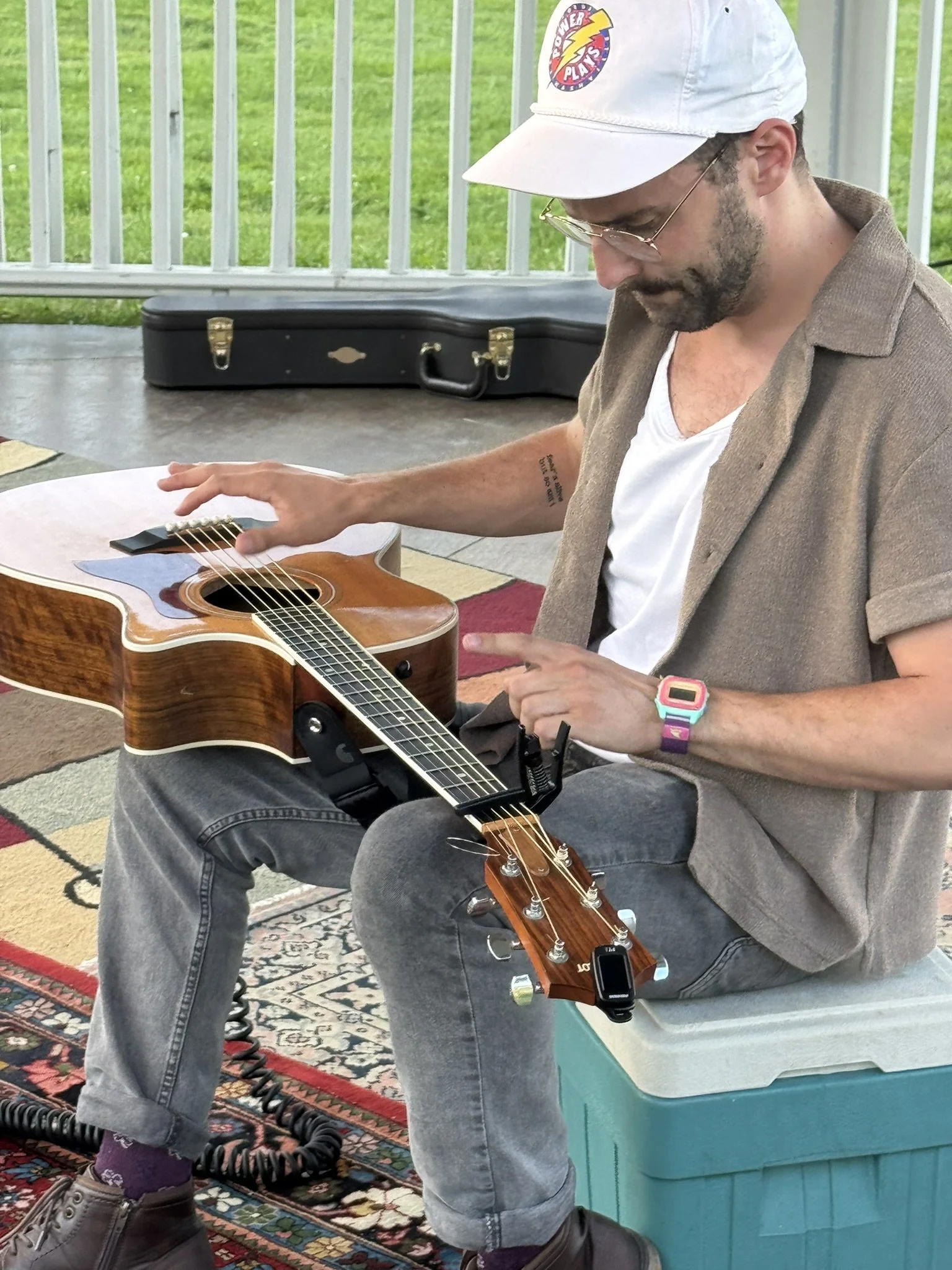 A man is sitting on a bench playing an acoustic guitar, with a guitar case in the background and decorative rug underneath. He is wearing a white cap, glasses, a brown shirt, and gray pants, with a colorful watch on his left wrist.