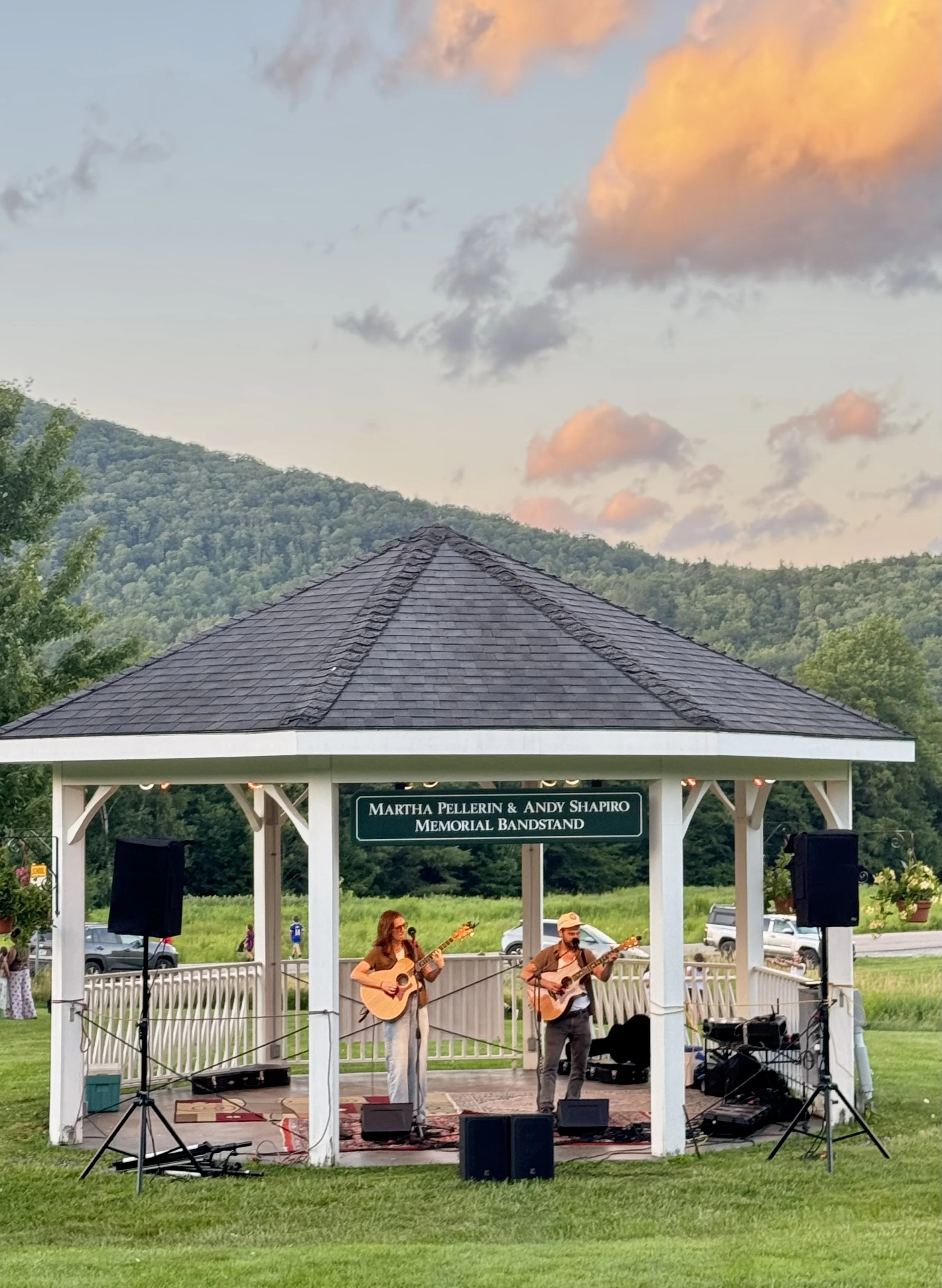 Musicians playing guitars on an outdoor stage in a pavilion with a sign reading "Martha Pellerin & Andy Shapiro Memorial Bandstand". The background features mountains and a sky with orange-tinted clouds at sunset.