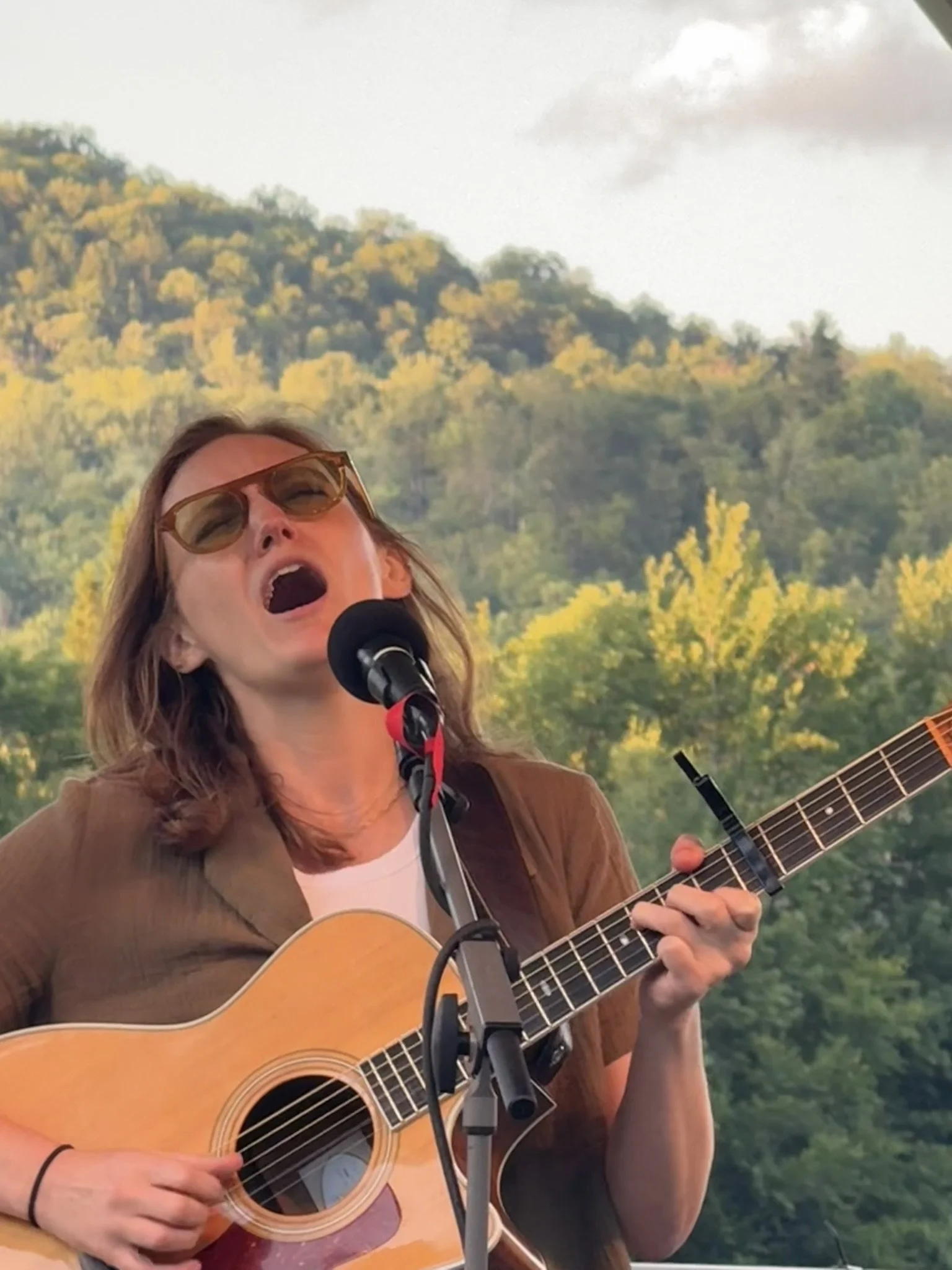 Woman with sunglasses singing into a microphone while playing an acoustic guitar outdoors, with green trees and hills in the background.