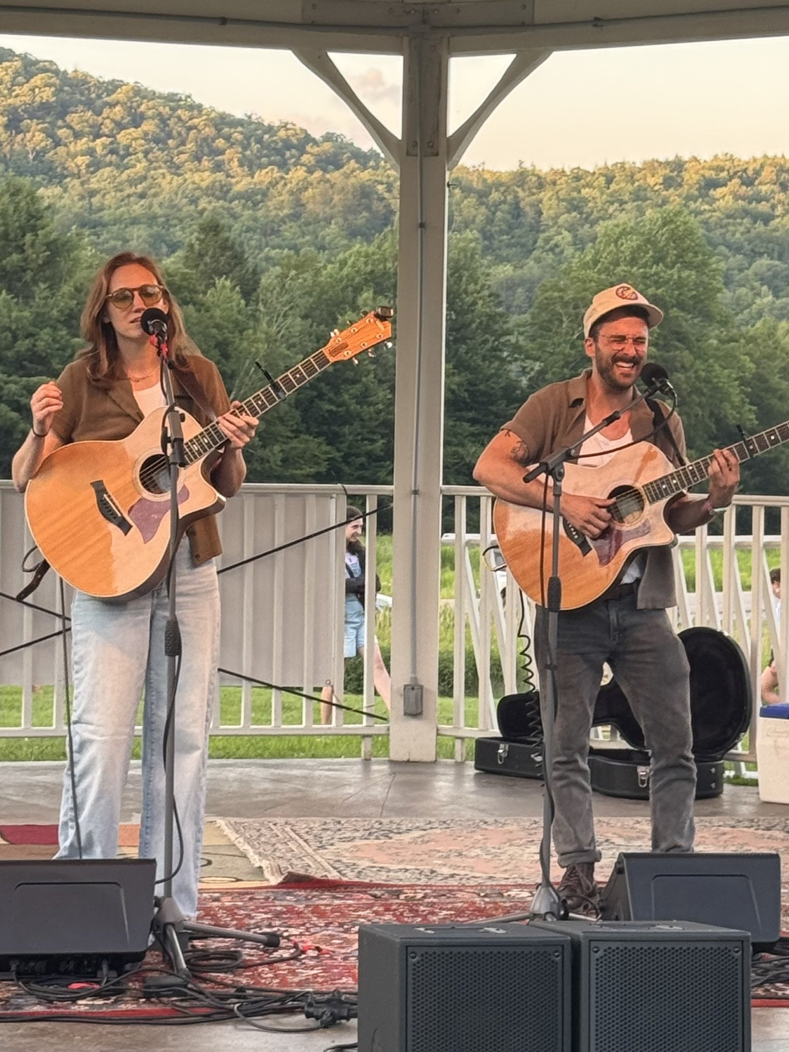 Two musicians performing with acoustic guitars on a stage outdoors, with a scenic background of trees and hills.