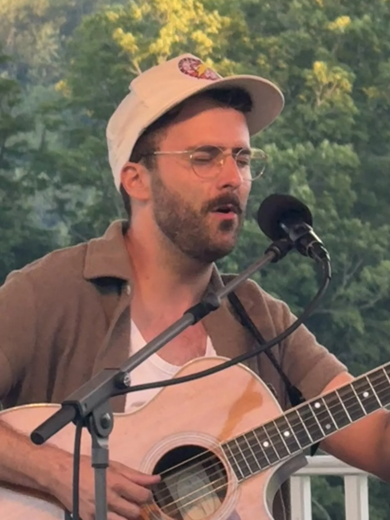 A man with glasses and a beard is playing an acoustic guitar and singing into a microphone outdoors with green trees in the background.