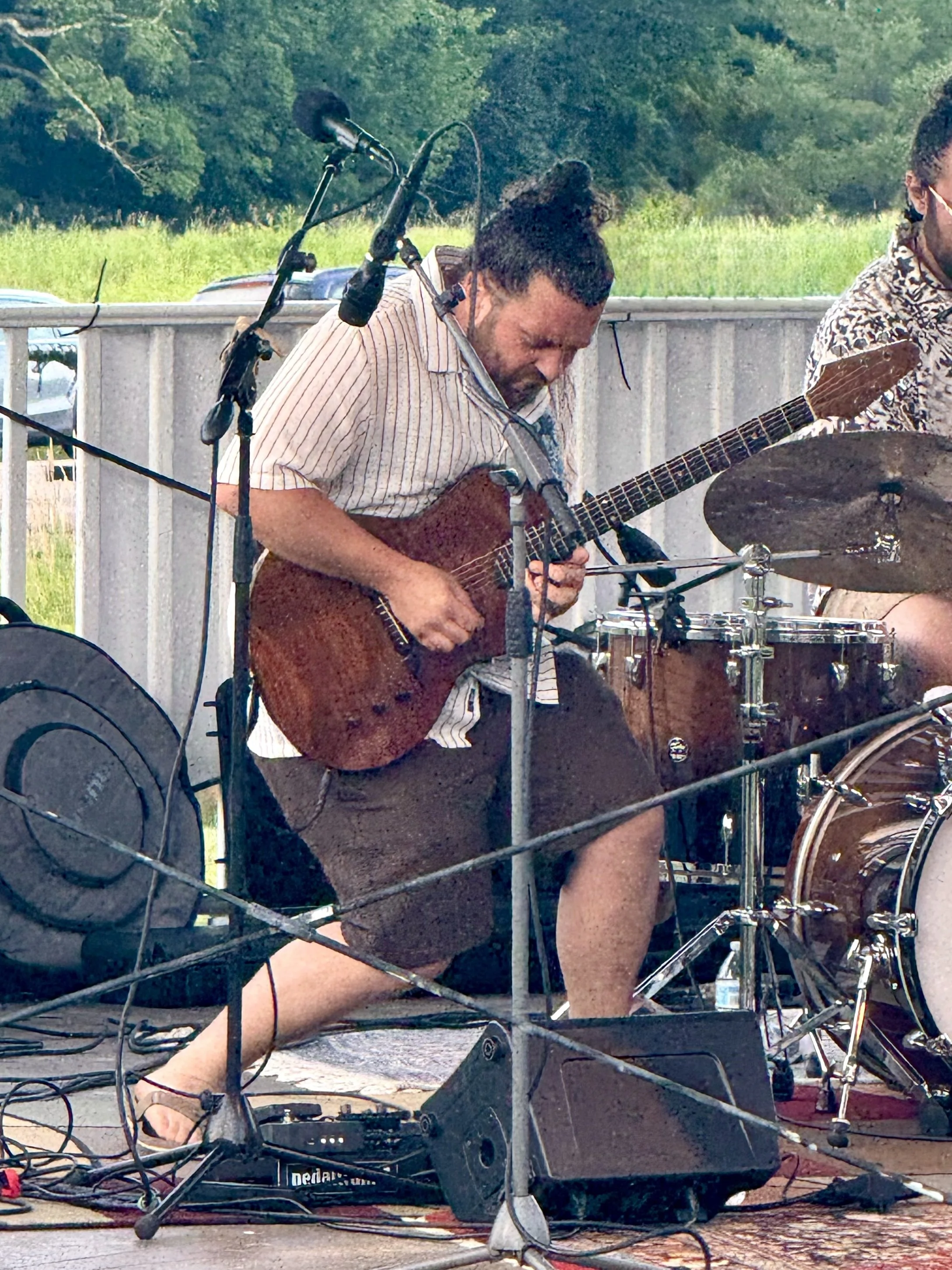 A person with dark curly hair tied up, wearing a striped shirt and shorts, playing an electric guitar on a stage outdoors with lush green trees in the background.