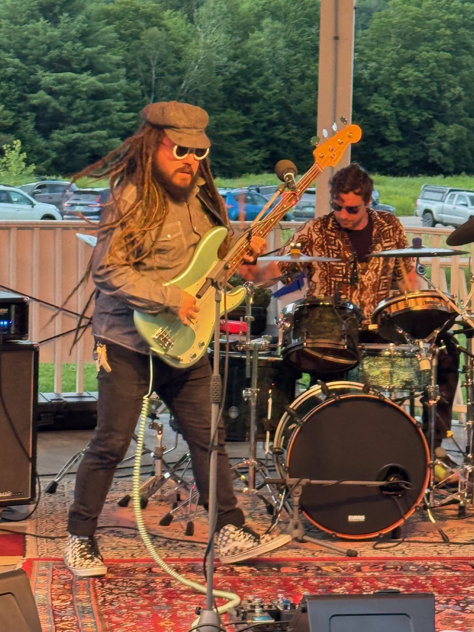 Two musicians performing outdoors on a stage, one playing an electric bass guitar and the other on drums. The stage has a patterned rug, and there are parked cars and green trees in the background.