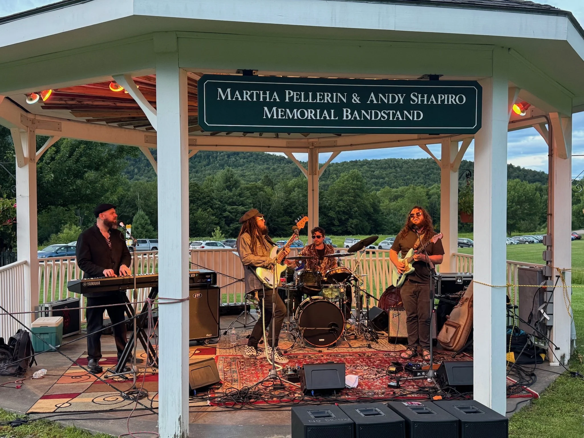 A band performing outdoors on a gazebo stage with a sign that reads 'Martha Pellerin & Andy Shapiro Memorial Bandstand.' The band has multiple musicians, including a keyboardist, a guitarist, a bassist, and a drummer, with a scenic green landscape an