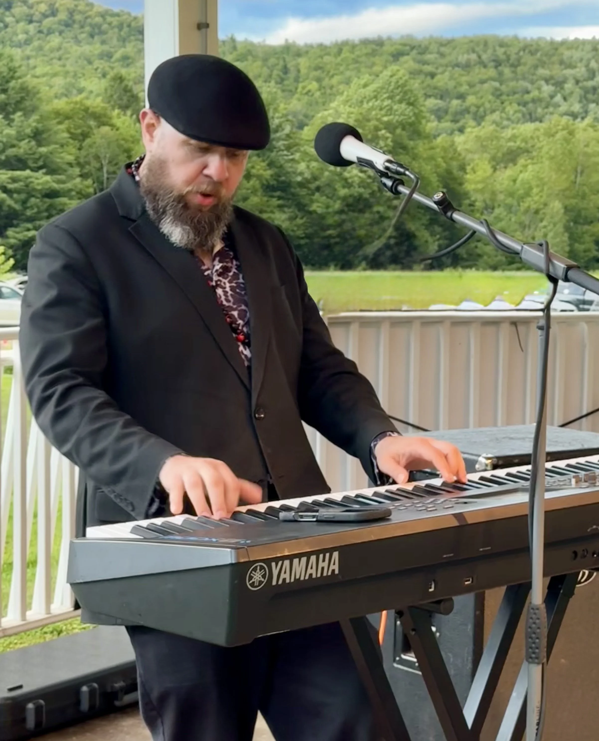 A man with a beard wearing a black blazer and leopard print shirt, playing a Yamaha keyboard at an outdoor event with green trees and blue sky in the background.