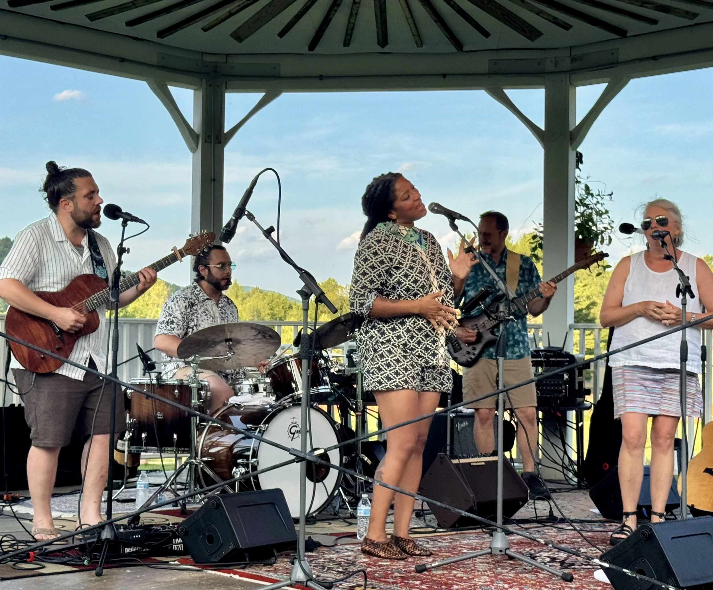 A band performing on an outdoor stage with a woman singing into a microphone, surrounded by musicians playing guitars and drums, under a pavilion with a scenic background of trees and sky.