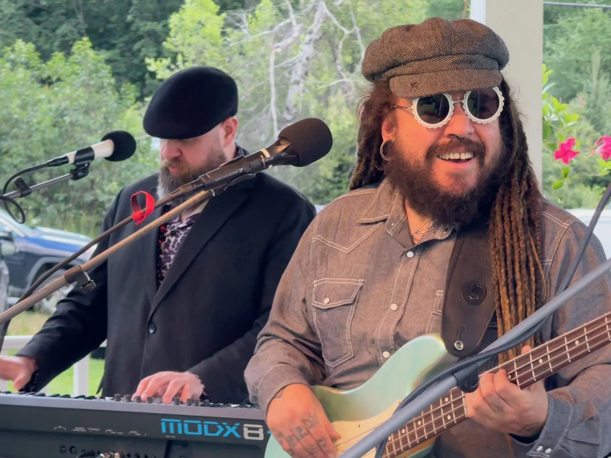 Two musicians performing outdoors, one playing keyboard and the other playing guitar, both wearing sunglasses and casual clothing with hats.