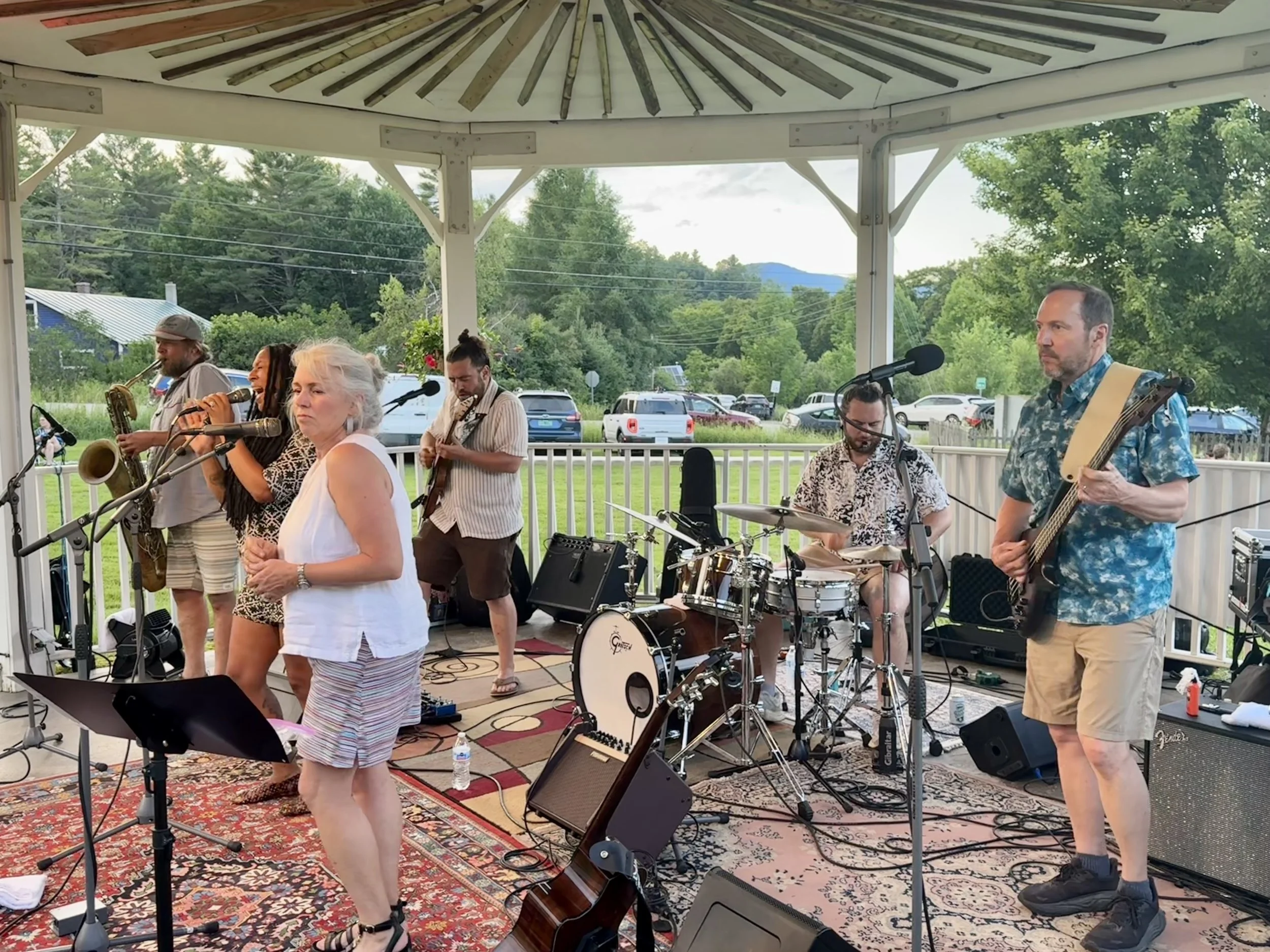 A band performing on a gazebo stage outdoors. The band includes a woman singing, a saxophonist, a guitarist, a drummer, and a bass player. The background shows trees, hills, and a parking lot filled with cars.