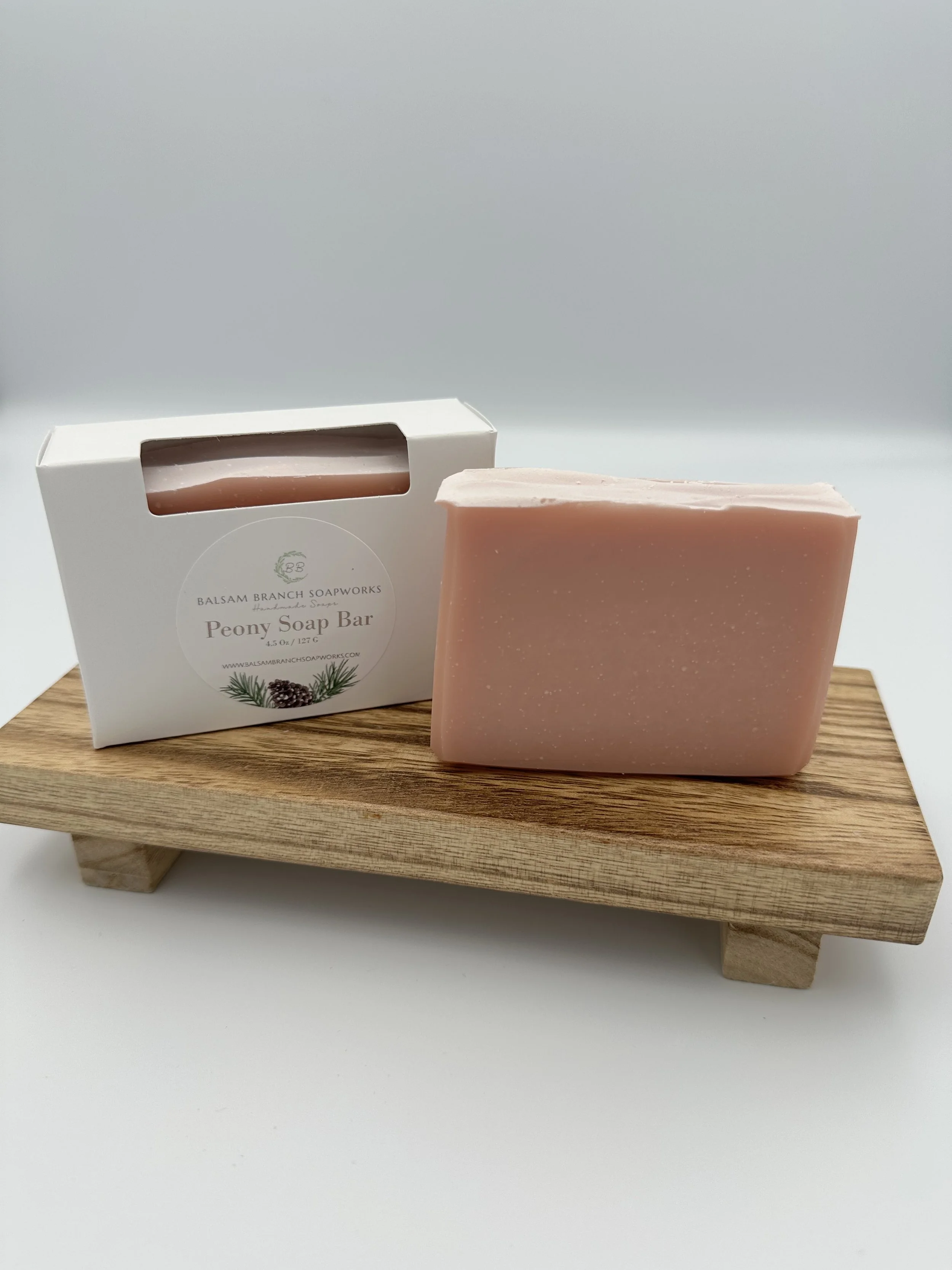 Pink rectangular bar of handmade soap next to packaging box labeled 'Peony Soap Bar' on a small wooden platform