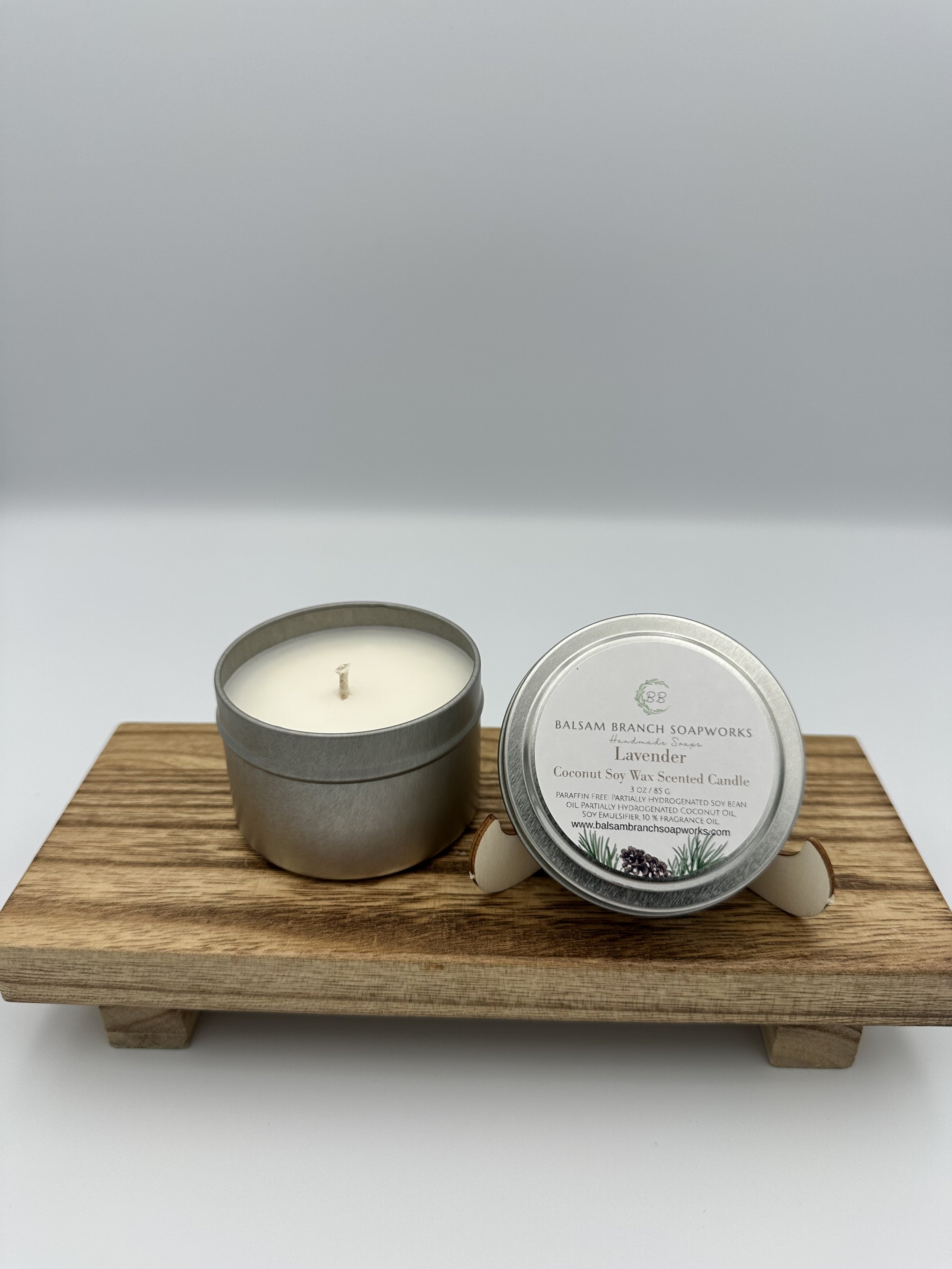 A scented lavender soy wax candle in a metal container and a matching tin candle on a wooden display tray.