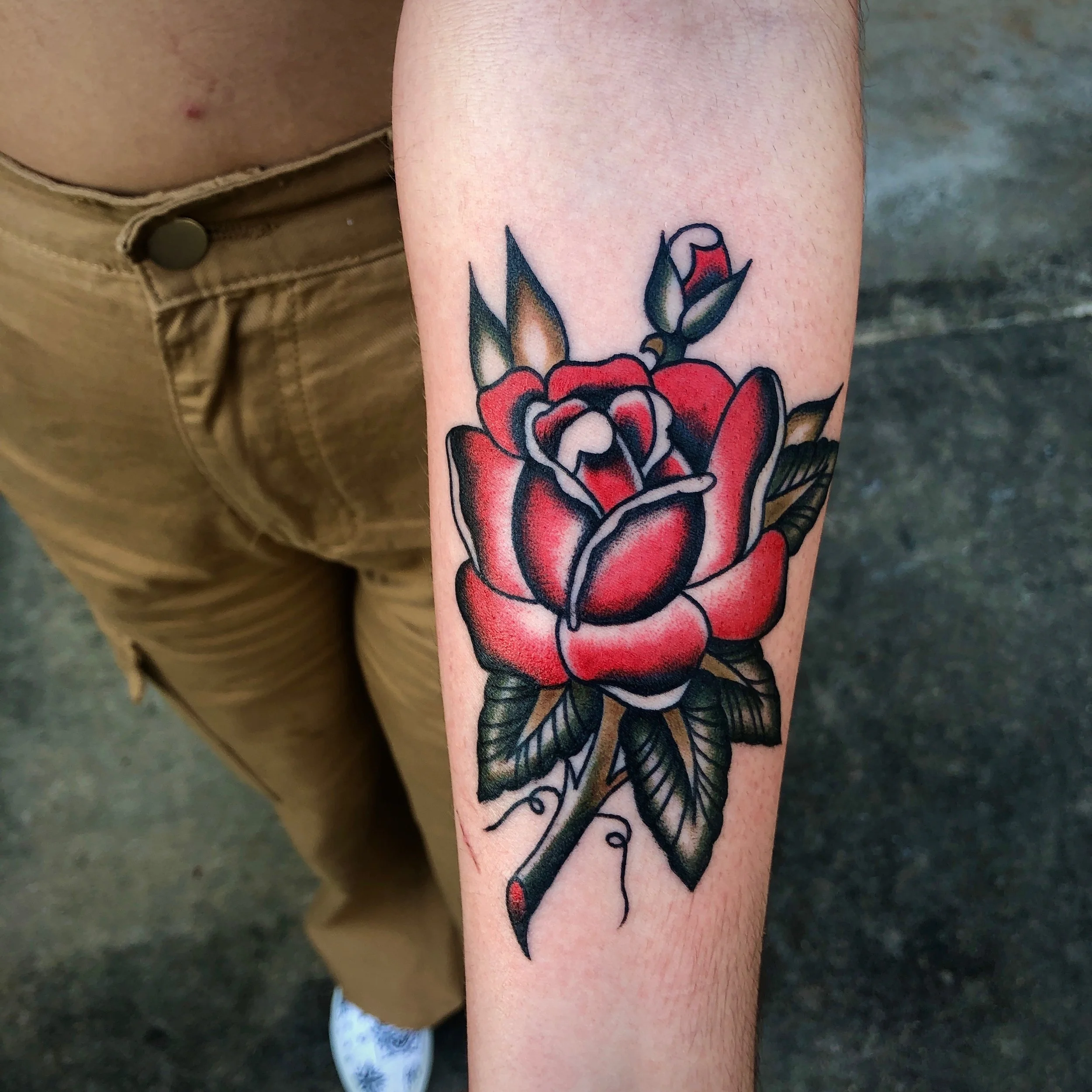 A colorful tattoo of a red rose with green leaves on a person's forearm.